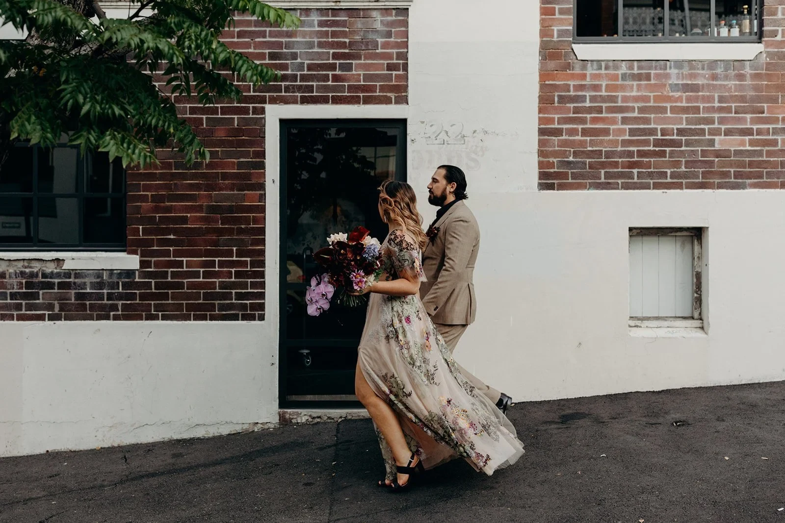 Victoria and Daniel walking past a brick building, with her floral skirt flowing and her bouquet by Mrs Gibbons Flowers, in Fortitude Valley, Brisbane, for an Elopement Collective elopement. Photo by Tessa Shannon.