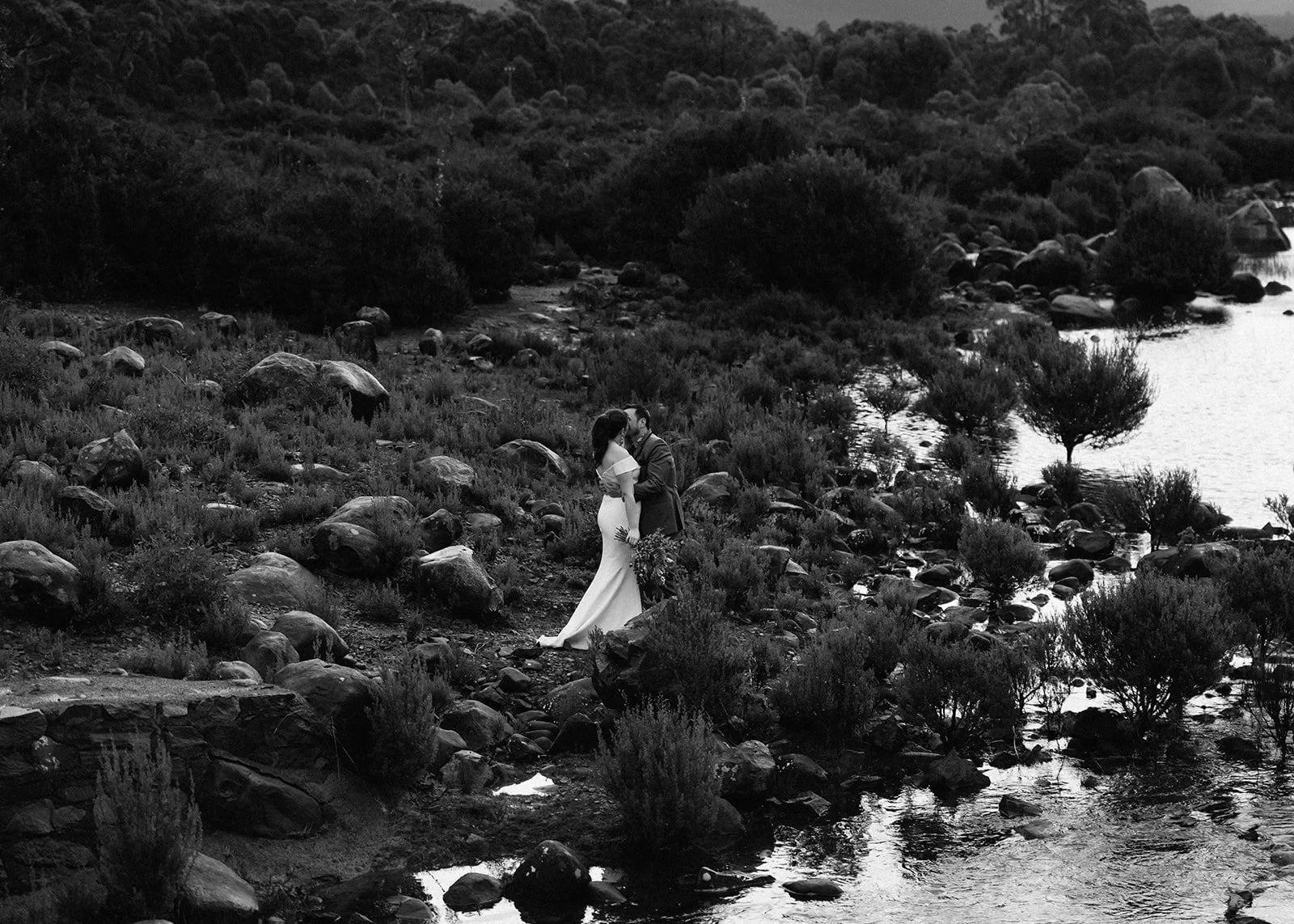 Black-and-white view from above of Sarah and Jono kissing on a rocky shore among tussock grasses, at Pumphouse Point, Lake St Clair, Tasmania, for an Elopement Collective elopement. Photo by Bulb Creative.