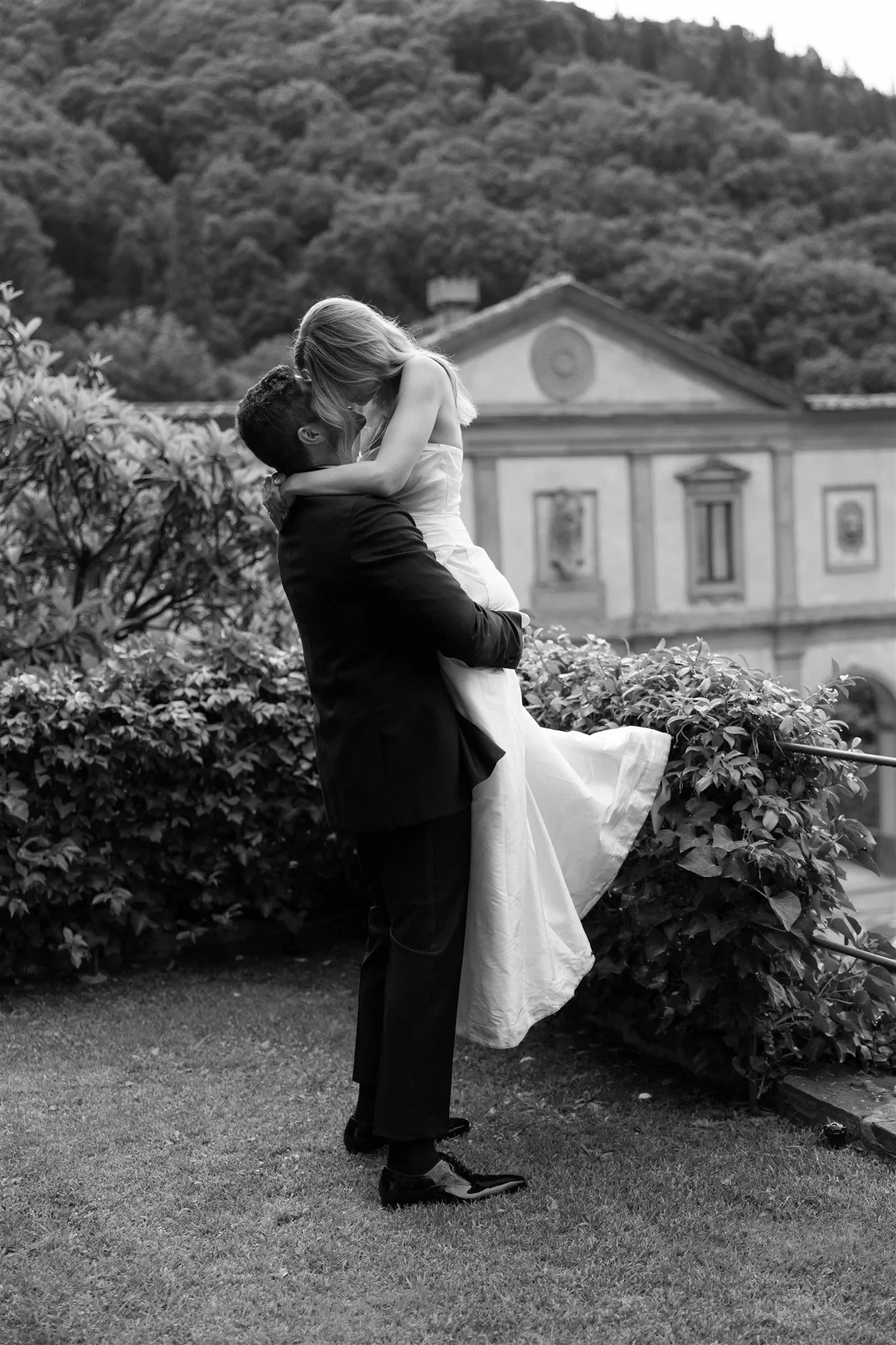 Black-and-white photograph of Matt carrying Holly through the garden in front of the villa, at Belmond Villa San Michele in Fiesole, overlooking Florence, Tuscany, Italy, for an Elopement Collective elopement. Photo by Khara from House of Lucie.