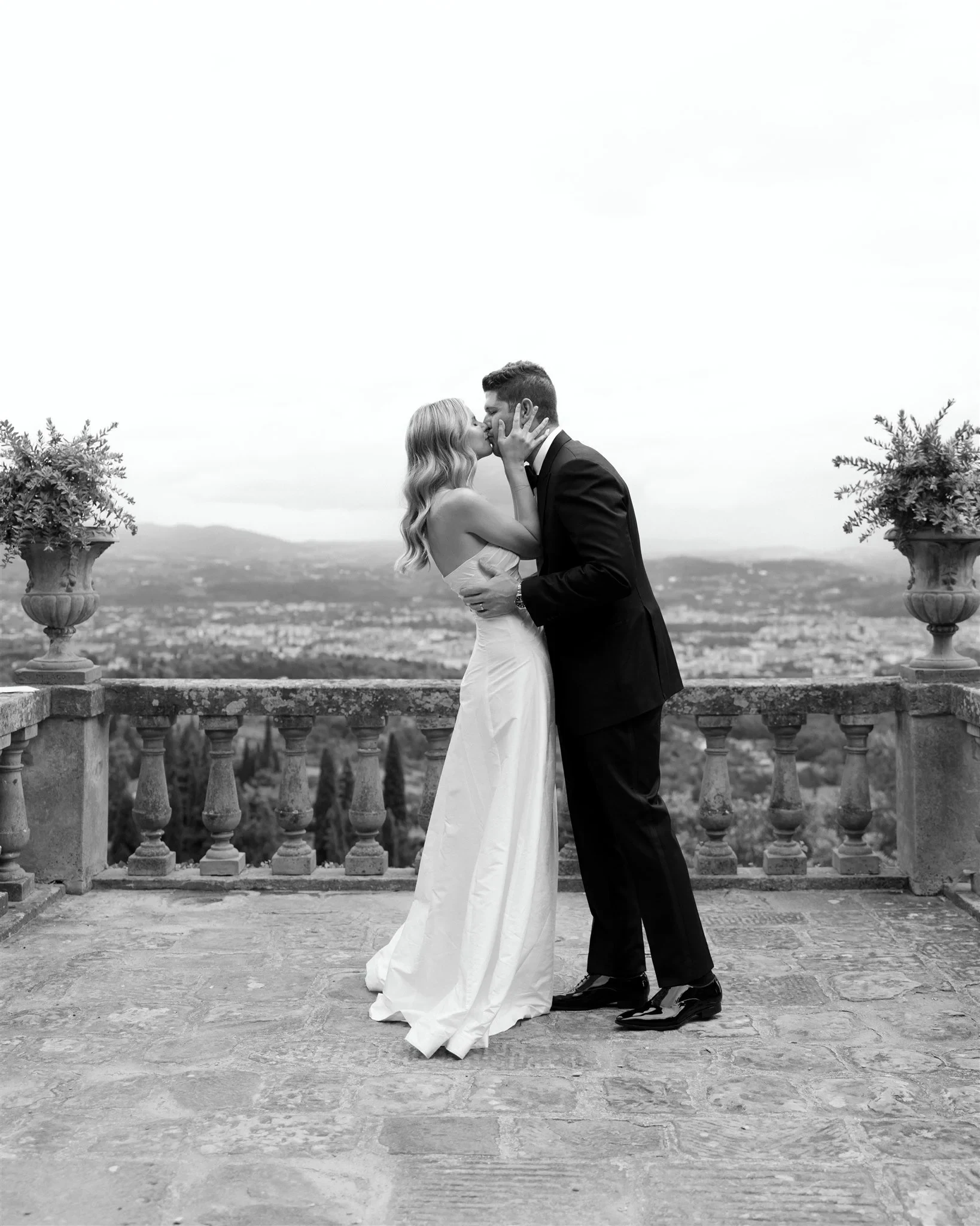 Black-and-white photograph of Holly and Matt kissing beside the terrace balustrade, at Belmond Villa San Michele in Fiesole, overlooking Florence, Tuscany, Italy, for an Elopement Collective elopement. Photo by Khara from House of Lucie.