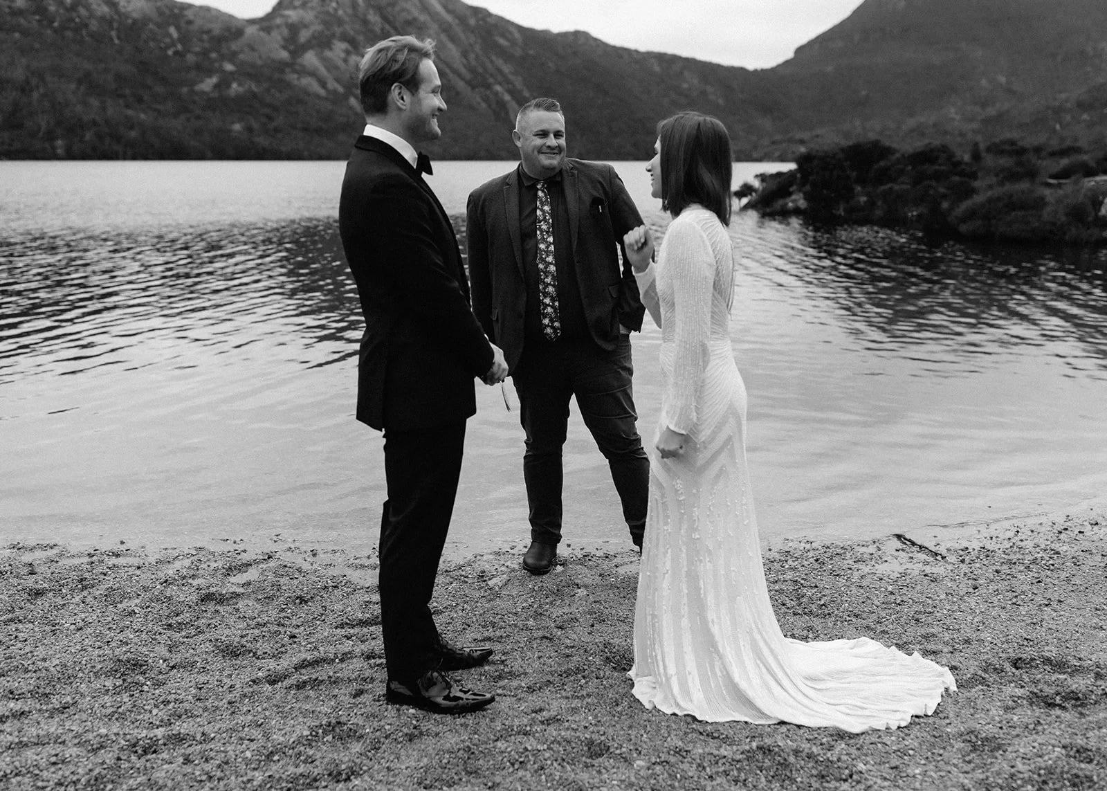 Black-and-white photograph of April and Josh facing one another with celebrant Josh Withers between them, at Dove Lake, Cradle Mountain, Tasmania, for an Elopement Collective elopement. Photo by Bulb Creative.