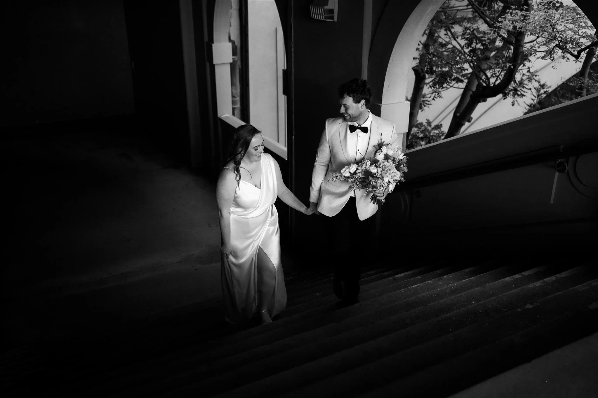 Black-and-white photograph of Alyce and Andrew holding hands as they climb stone steps, in The Rocks, Sydney, New South Wales, for an Elopement Collective elopement. Photo by Jason Corroto.