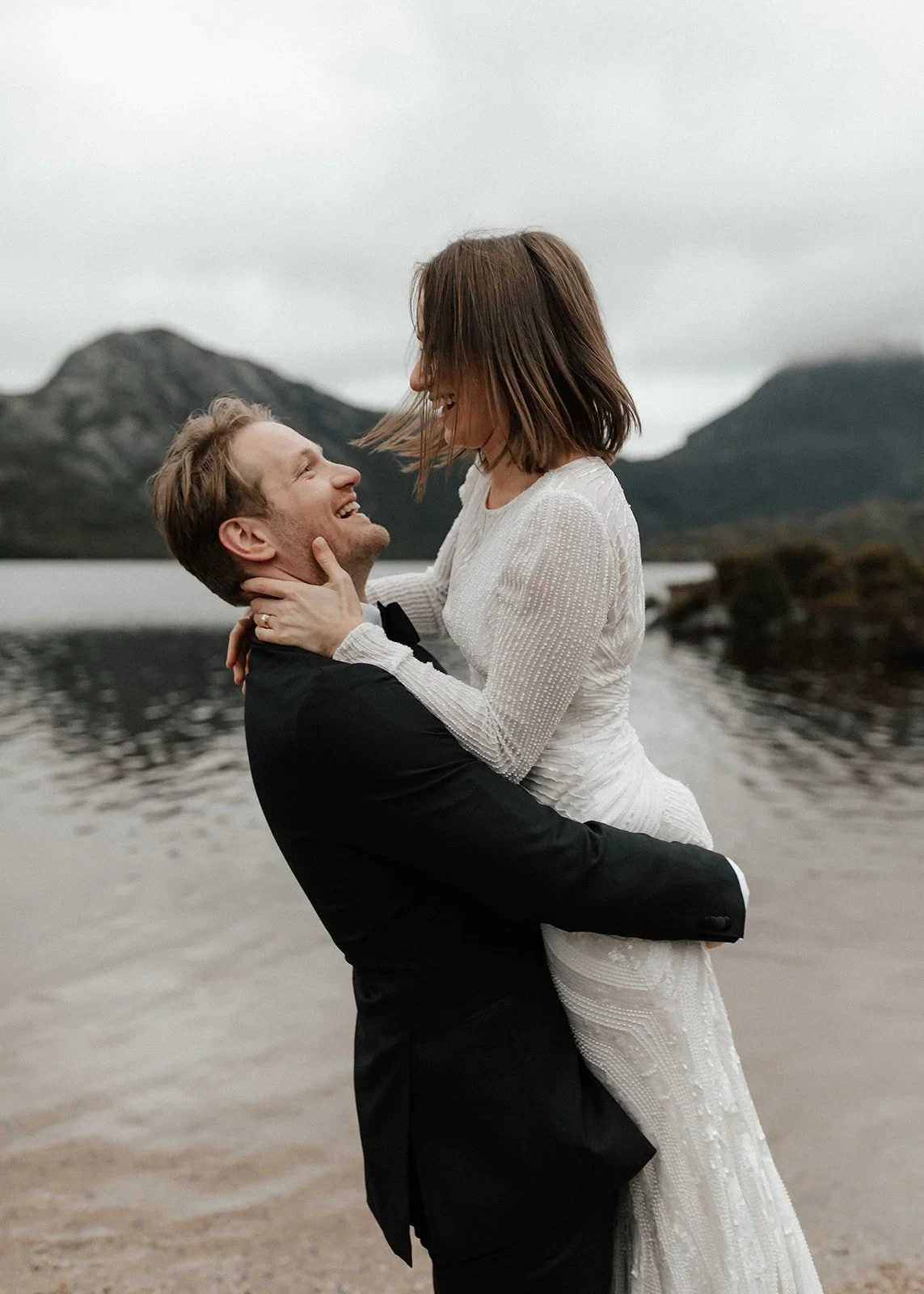 Josh lifting April as they laugh together beside the lake, at Dove Lake, Cradle Mountain, Tasmania, for an Elopement Collective elopement. Photo by Bulb Creative.