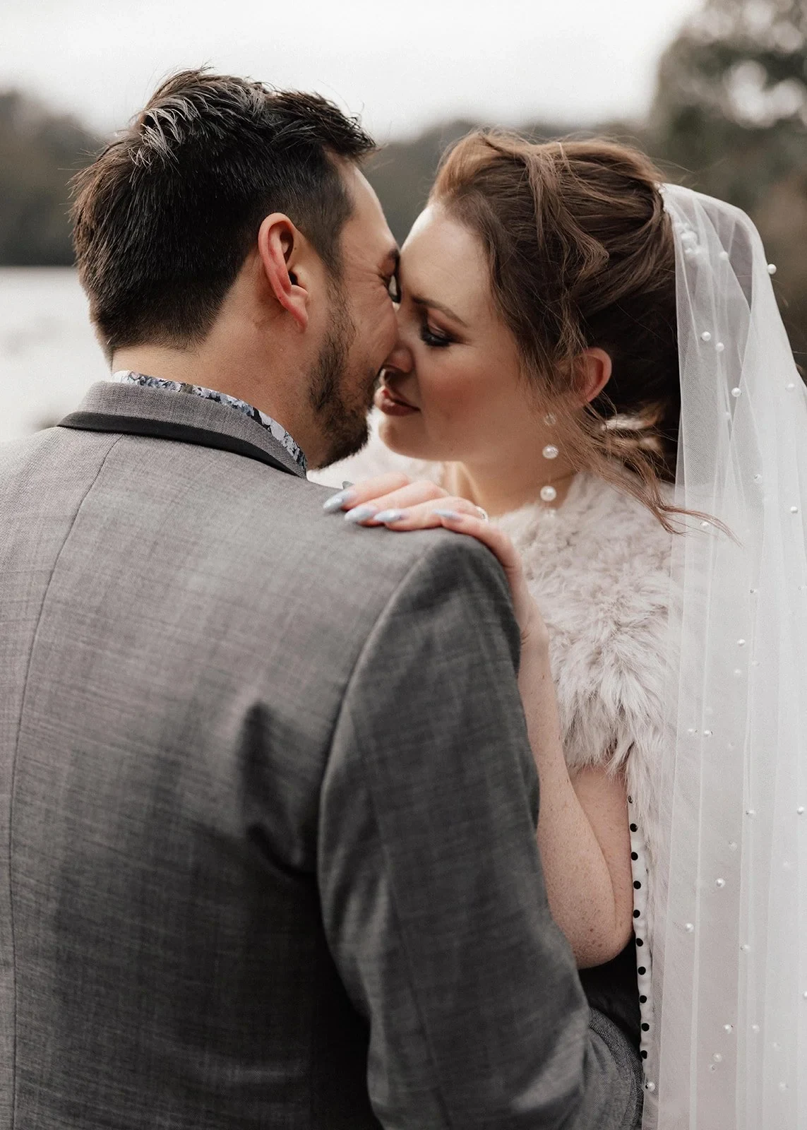 Sarah and Jono touching foreheads beside the lake, with her pale textured wrap around her shoulders, at Pumphouse Point, Lake St Clair, Tasmania, for an Elopement Collective elopement. Photo by Bulb Creative.