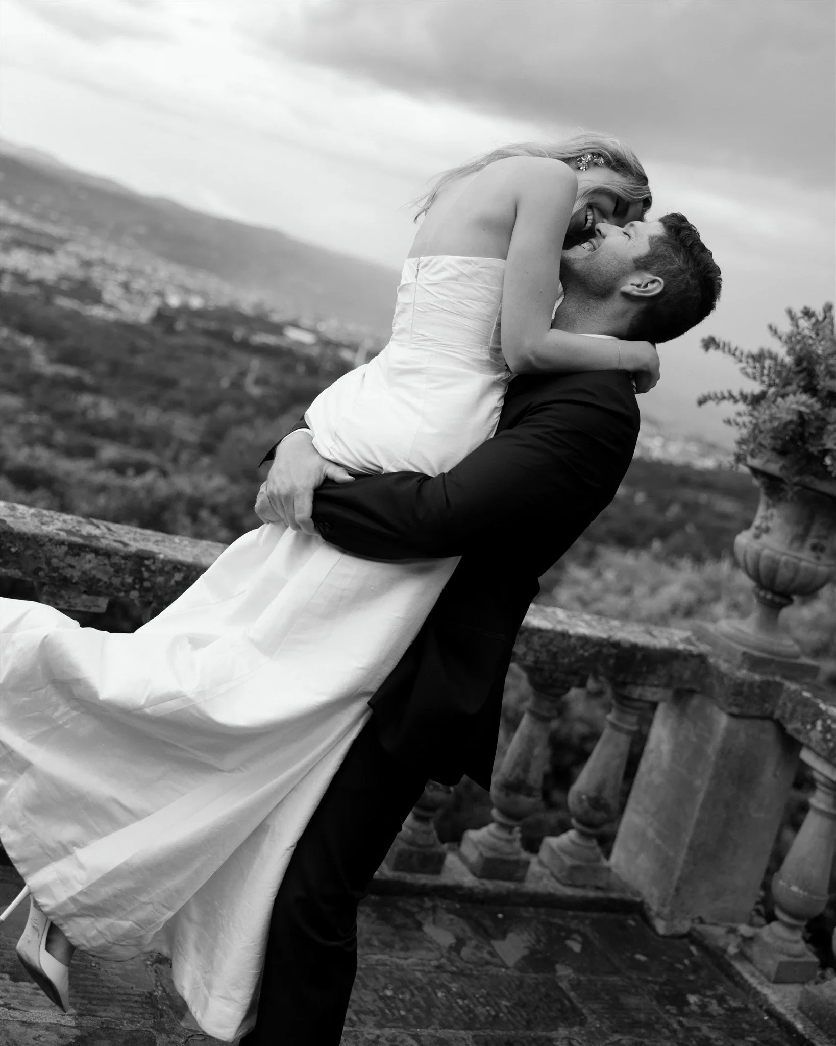 Black-and-white photograph of Matt lifting Holly into a kiss beside a terrace overlooking Florence, at Belmond Villa San Michele in Fiesole, overlooking Florence, Tuscany, Italy, for an Elopement Collective elopement. Photo by Khara from House of Lucie.