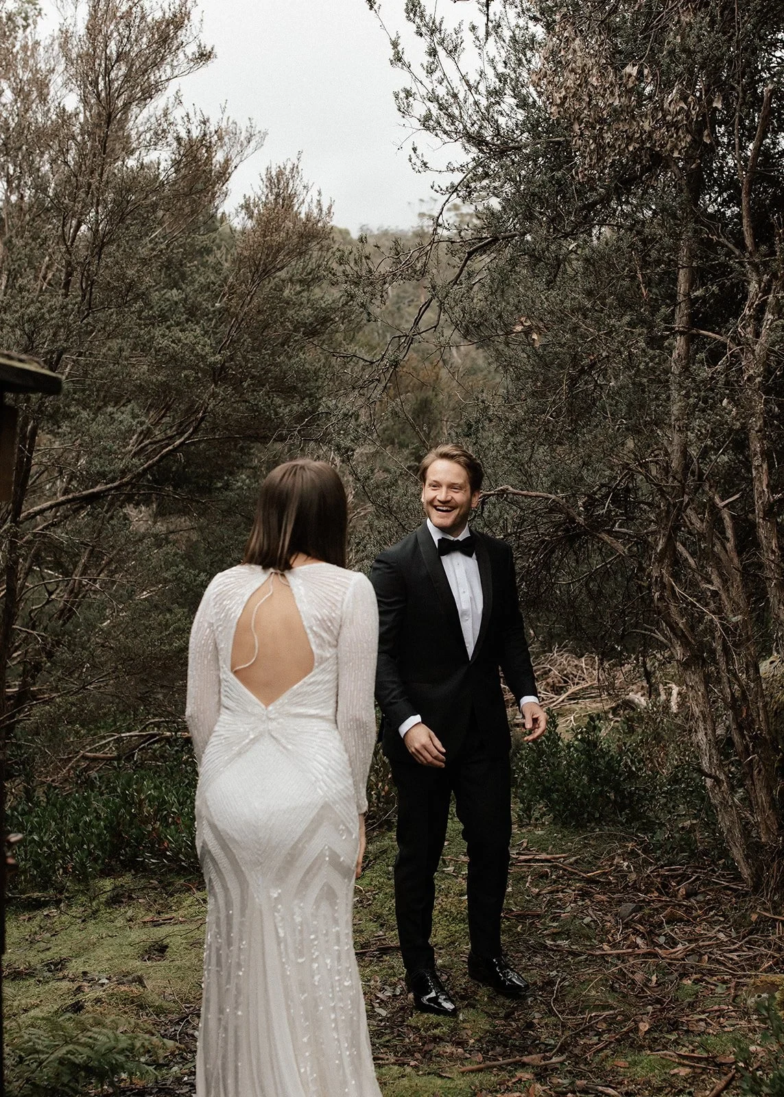 April approaching Josh along a mossy woodland path as he smiles at her, at Cradle Mountain Lodge in Cradle Mountain, Tasmania, for an Elopement Collective elopement. Photo by Bulb Creative.