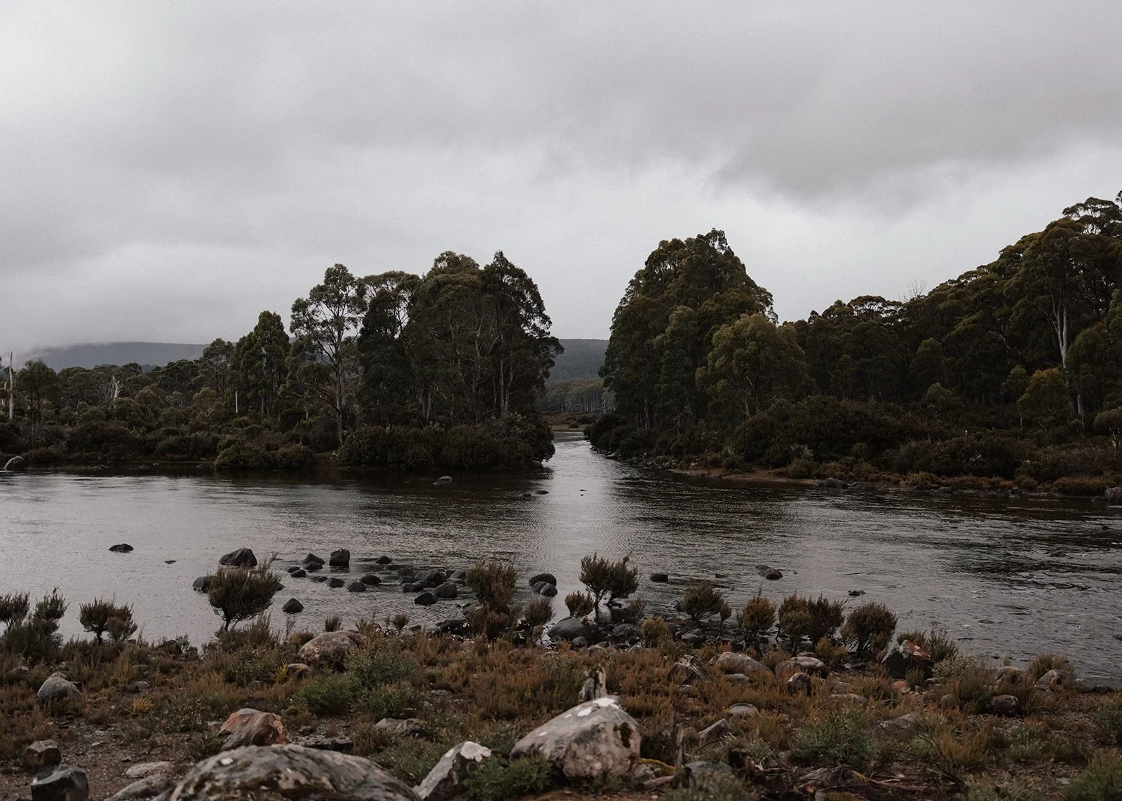 Wide view of Sarah and Jono kissing beside the lake, surrounded by rocks, bush and still water, at Pumphouse Point, Lake St Clair, Tasmania, for an Elopement Collective elopement. Photo by Bulb Creative.