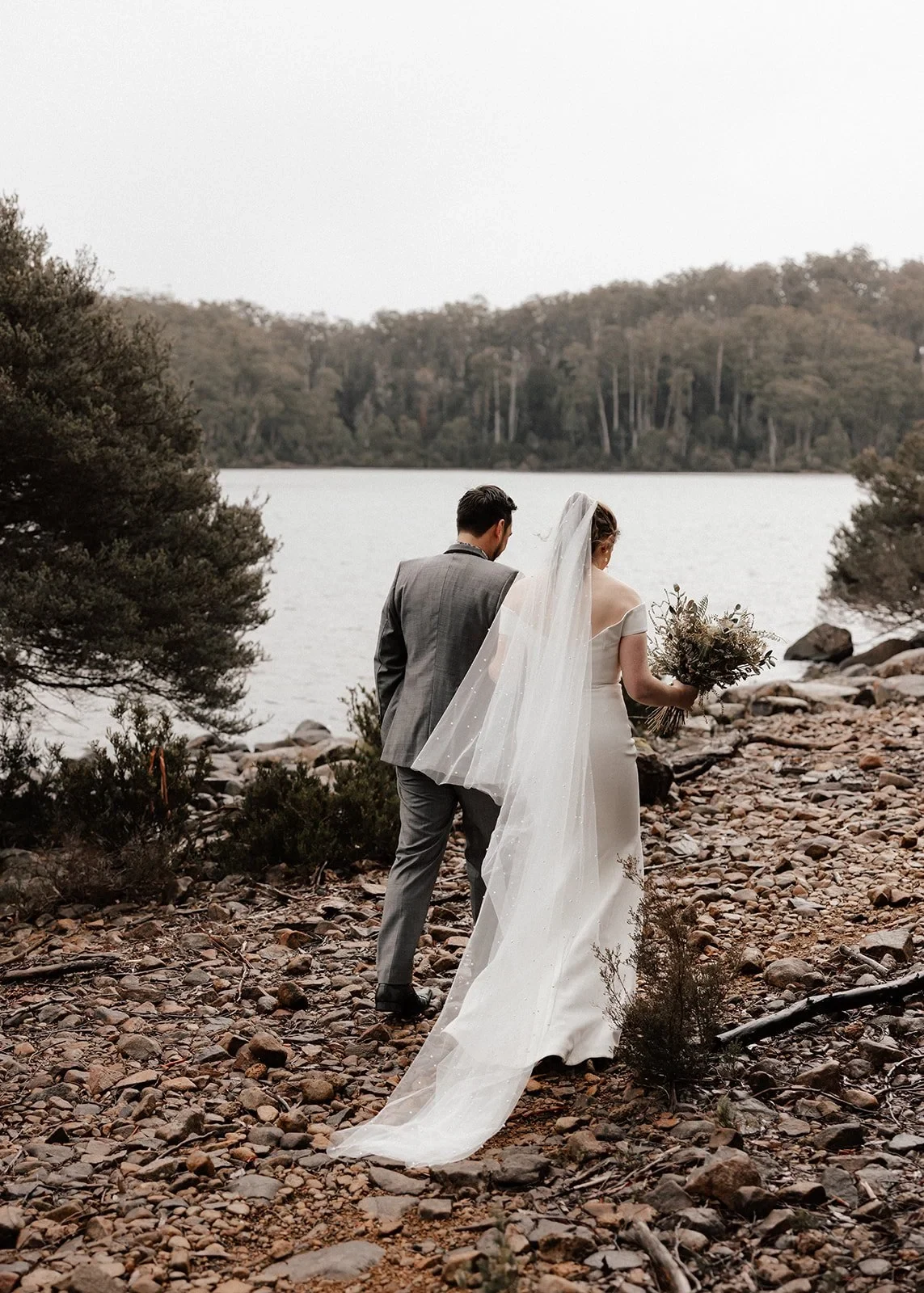 Sarah and Jono walking hand in hand towards the lake, her long veil trailing behind them, at Pumphouse Point, Lake St Clair, Tasmania, for an Elopement Collective elopement. Photo by Bulb Creative.