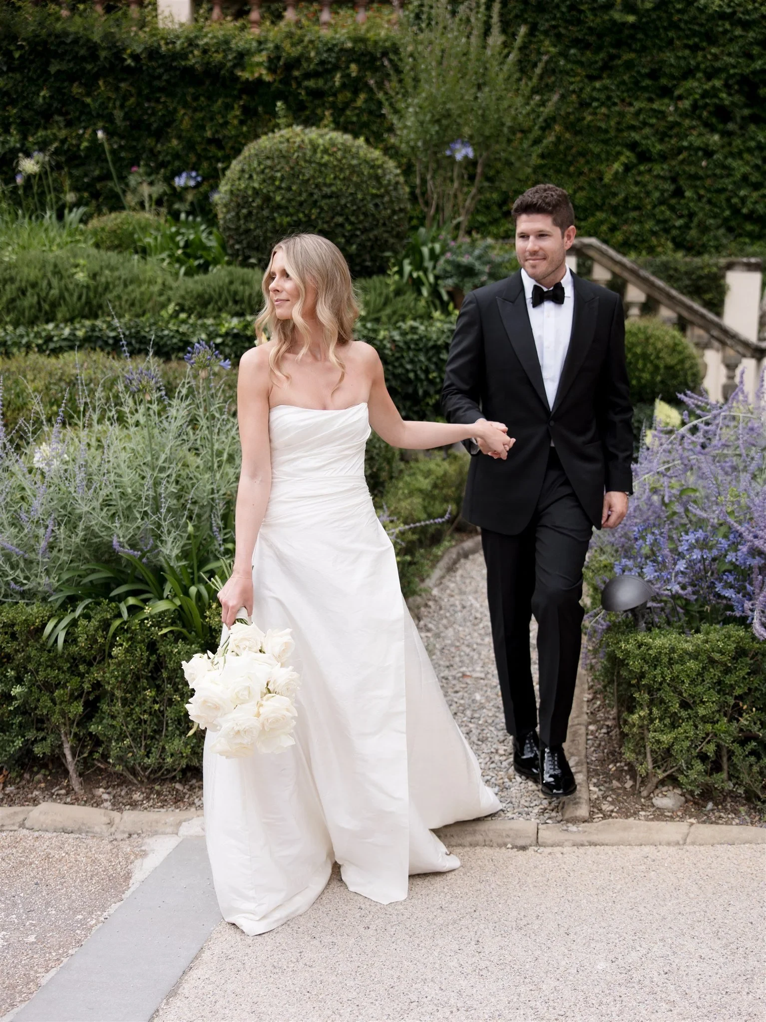 Holly leading Matt through purple garden flowers, carrying her white bouquet by Le Francesche, at Belmond Villa San Michele in Fiesole, overlooking Florence, Tuscany, Italy, for an Elopement Collective elopement. Photo by Khara from House of Lucie.