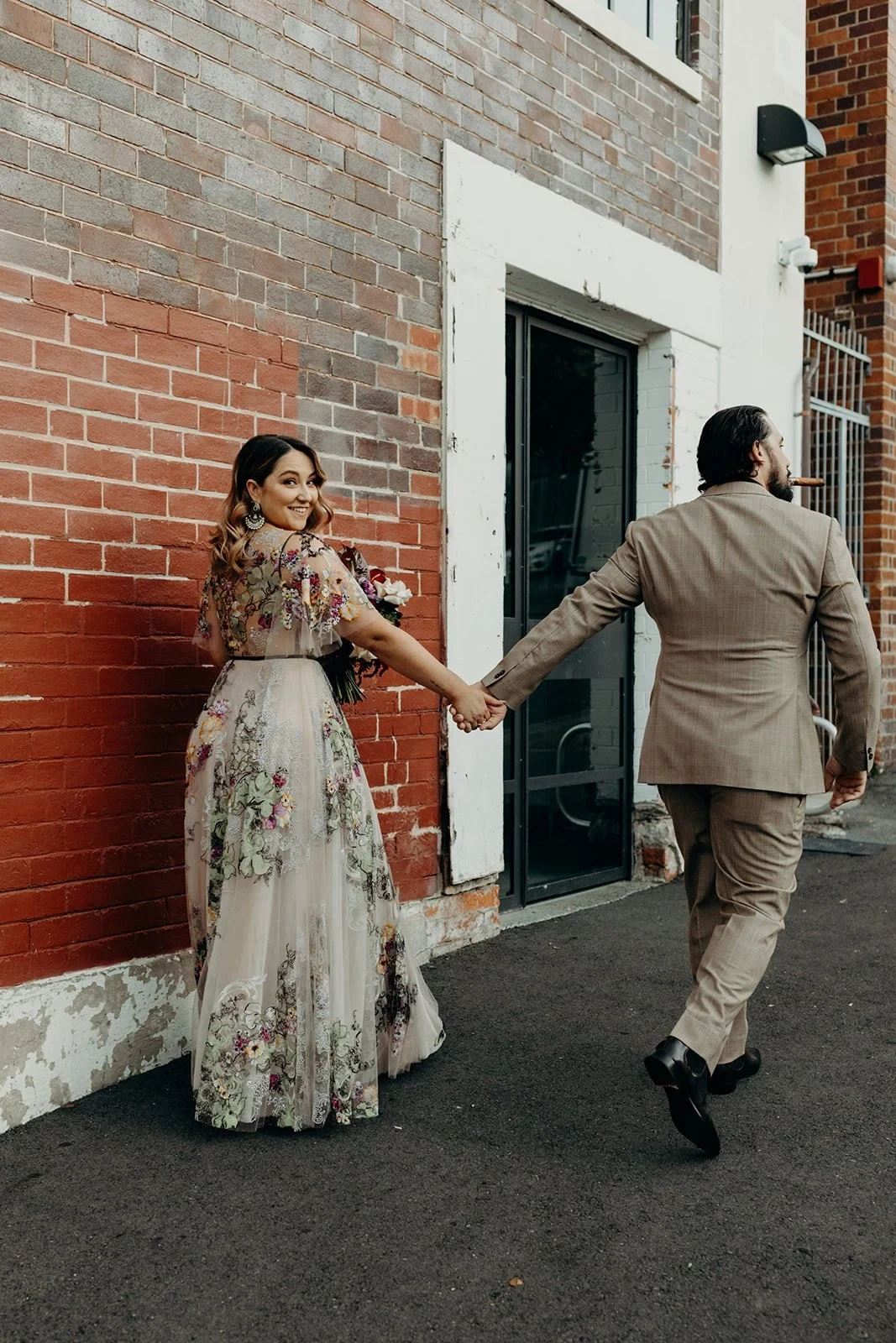 Daniel stepping away while holding Victoria’s hand beside a red brick building, in Fortitude Valley, Brisbane, for an Elopement Collective elopement. Photo by Tessa Shannon.