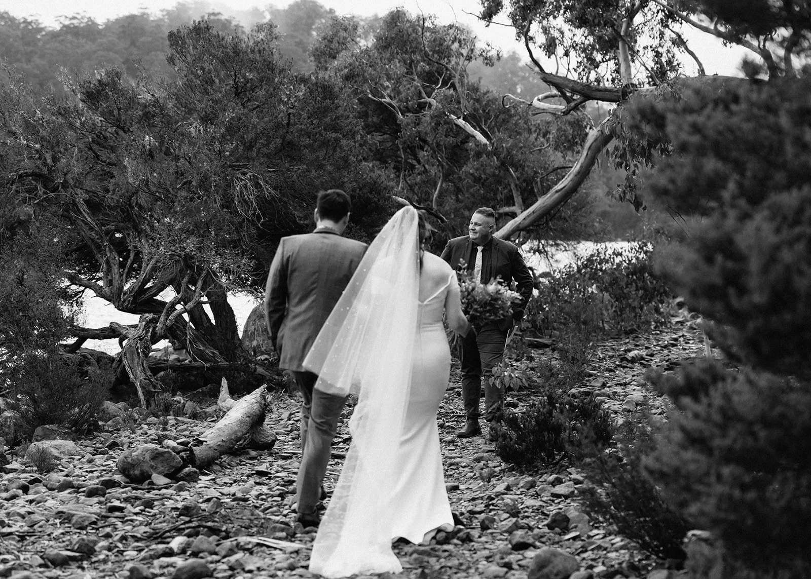 Black-and-white photograph of Sarah and Jono approaching celebrant Josh Withers along a rocky lakeside path, at Pumphouse Point, Lake St Clair, Tasmania, for an Elopement Collective elopement. Photo by Bulb Creative.