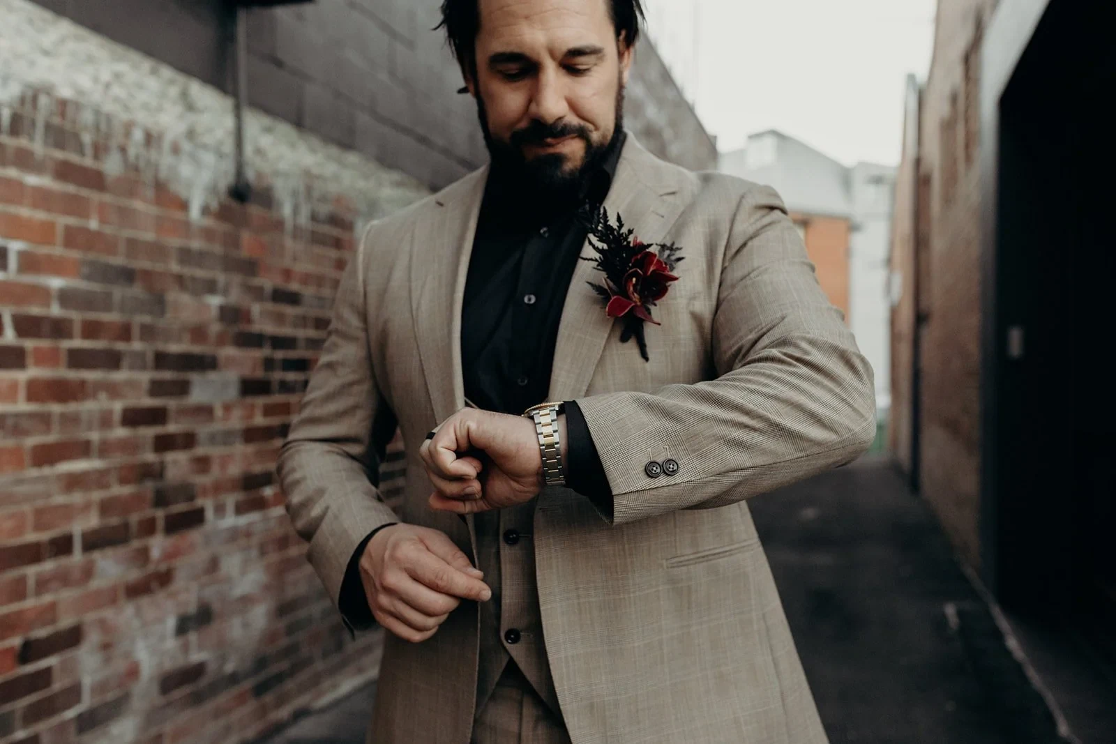 Daniel adjusting his wristwatch, with his beige suit and burgundy floral buttonhole visible, in Fortitude Valley, Brisbane, for an Elopement Collective elopement. Photo by Tessa Shannon.