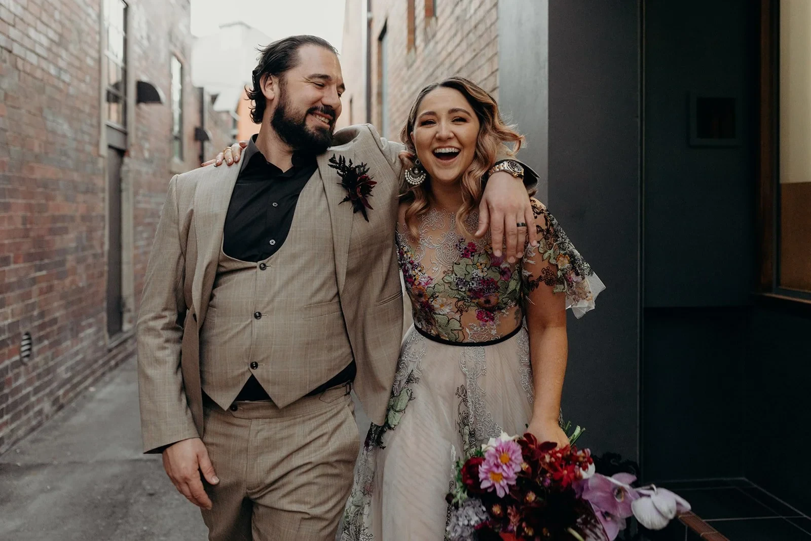 Victoria and Daniel laughing as they walk arm in arm along a brick-lined laneway, with her bouquet by Mrs Gibbons Flowers, in Fortitude Valley, Brisbane, for an Elopement Collective elopement. Photo by Tessa Shannon.
