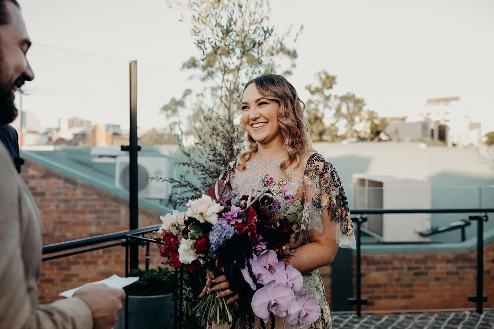 Victoria smiling at Daniel while holding her colourful bouquet by Mrs Gibbons Flowers during their ceremony, at Agnes Restaurant in Fortitude Valley, Brisbane, for an Elopement Collective elopement. Photo by Tessa Shannon.