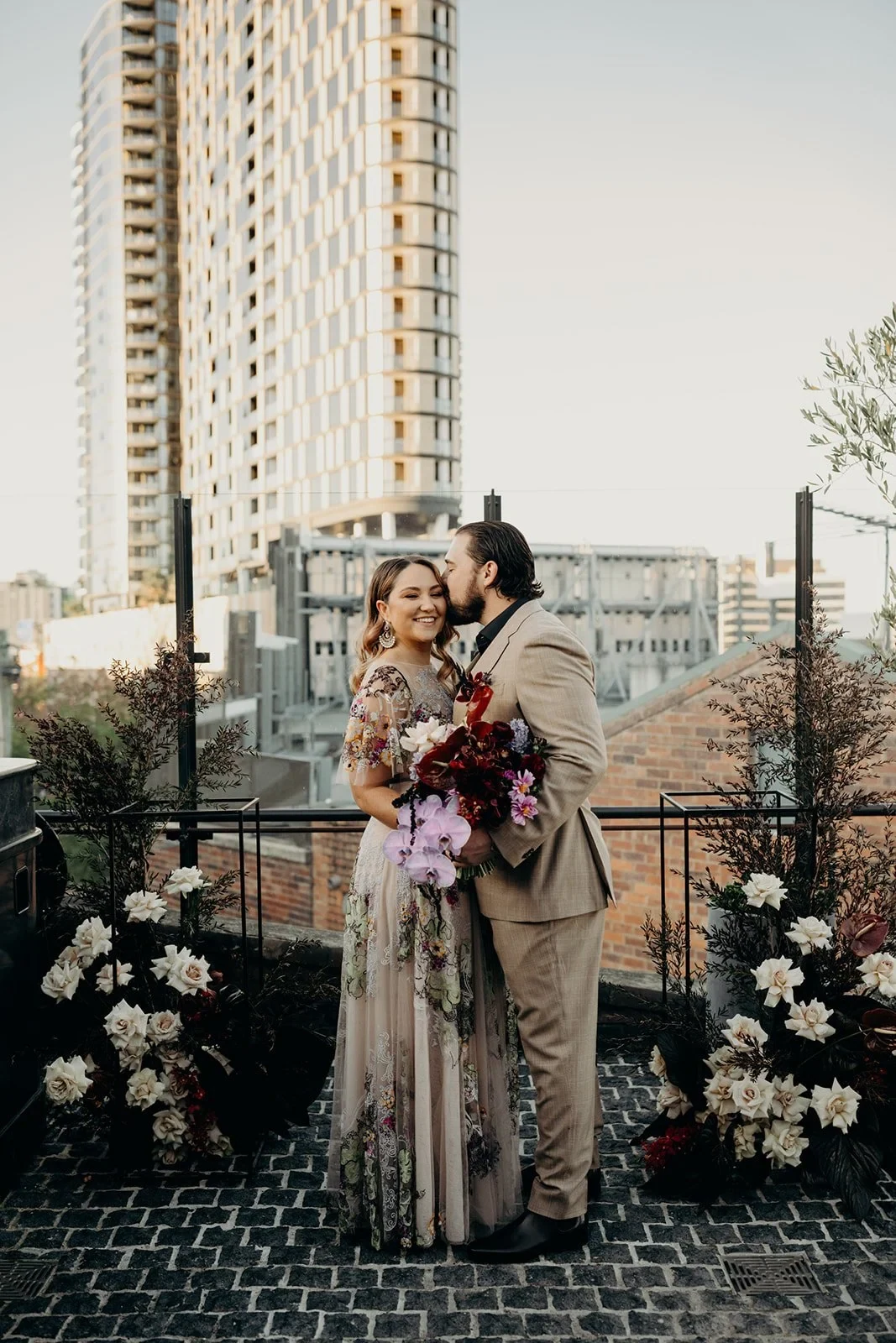 Victoria smiling in Daniel’s embrace among white rooftop flowers by Mrs Gibbons Flowers, at Agnes Restaurant in Fortitude Valley, Brisbane, for an Elopement Collective elopement. Photo by Tessa Shannon.
