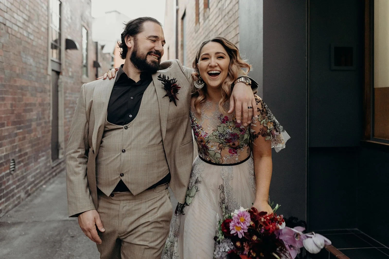 Victoria and Daniel laughing as they walk arm in arm along a brick-lined laneway, with her bouquet by Mrs Gibbons Flowers, in Fortitude Valley, Brisbane, for an Elopement Collective elopement. Photo by Tessa Shannon.