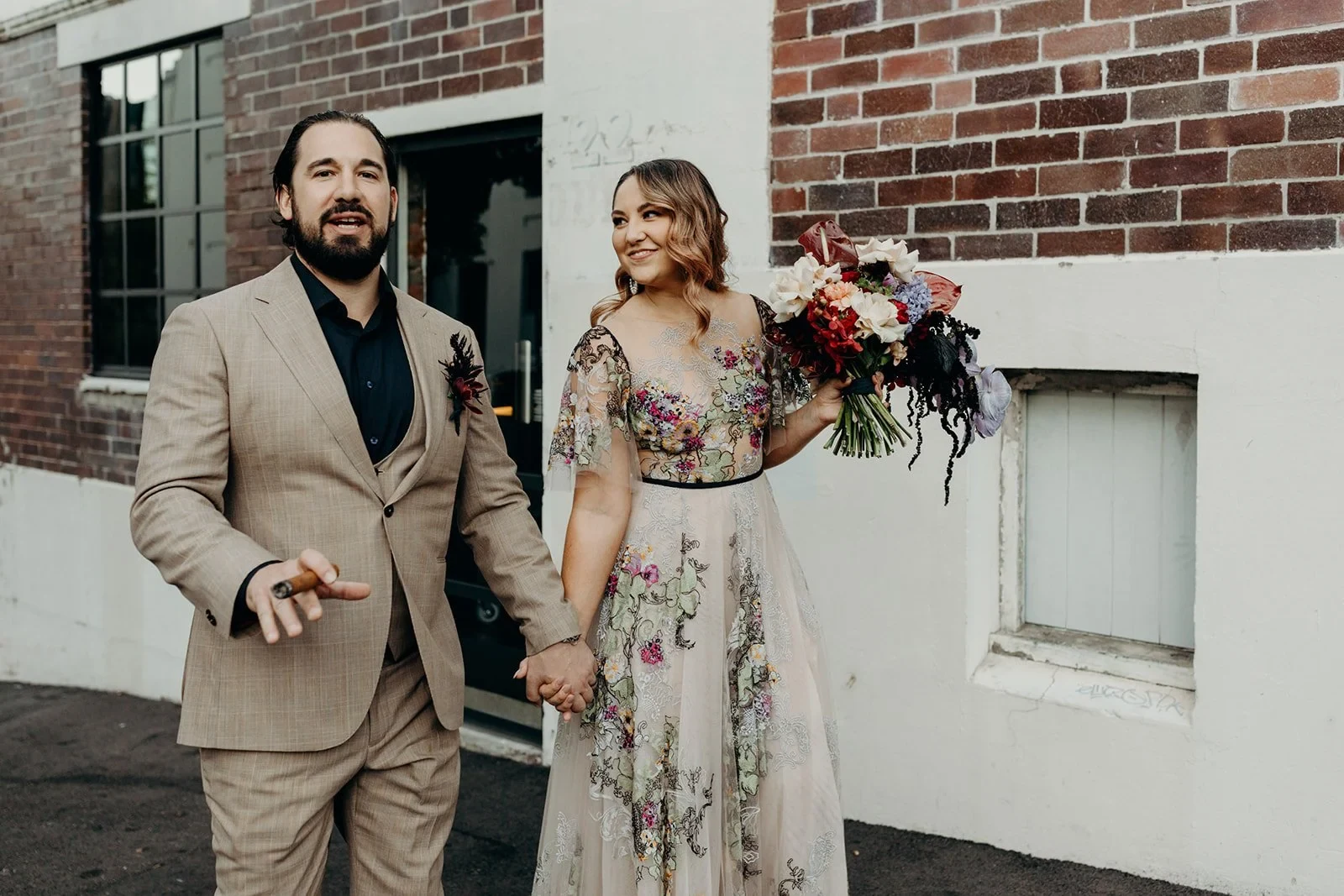 Victoria and Daniel standing outside a brick building, with her colourful bouquet by Mrs Gibbons Flowers, in Fortitude Valley, Brisbane, for an Elopement Collective elopement. Photo by Tessa Shannon.