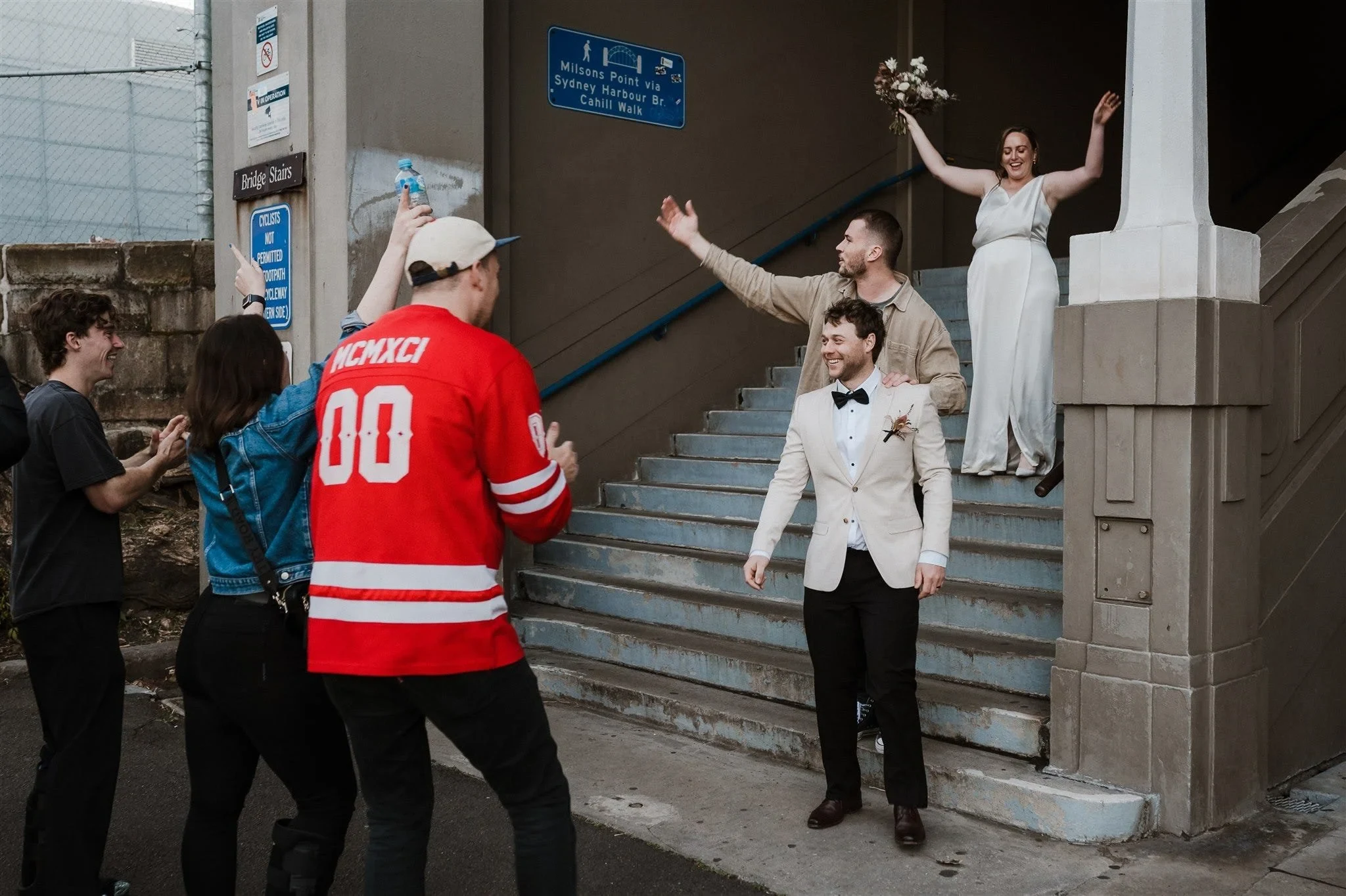 Alyce and Andrew raising their arms on stone steps while guests photograph and film them, in The Rocks, Sydney, New South Wales, for an Elopement Collective elopement. Photo by Jason Corroto.