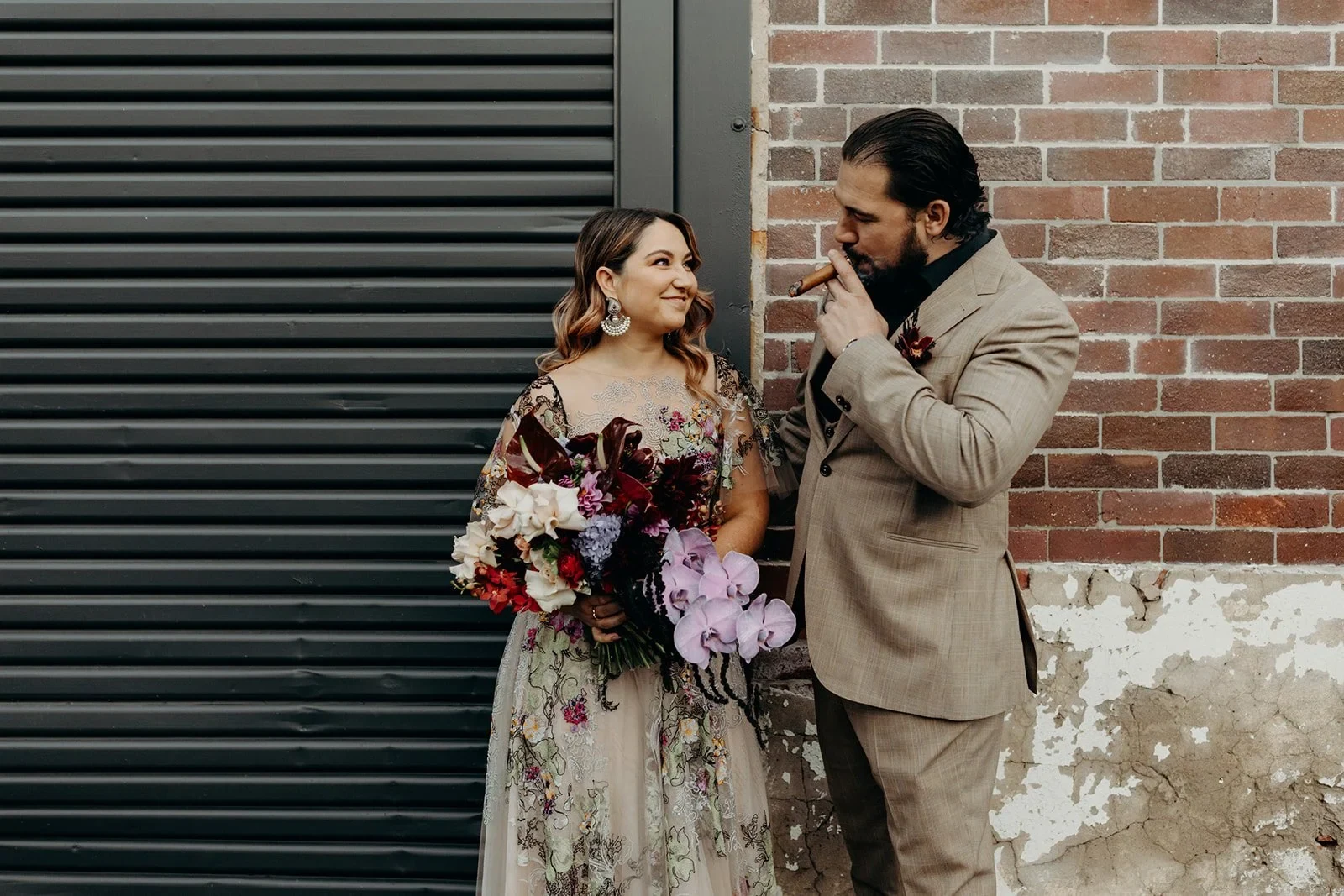 Victoria smiling at Daniel as he smokes a cigar beside her bouquet by Mrs Gibbons Flowers, in Fortitude Valley, Brisbane, for an Elopement Collective elopement. Photo by Tessa Shannon.