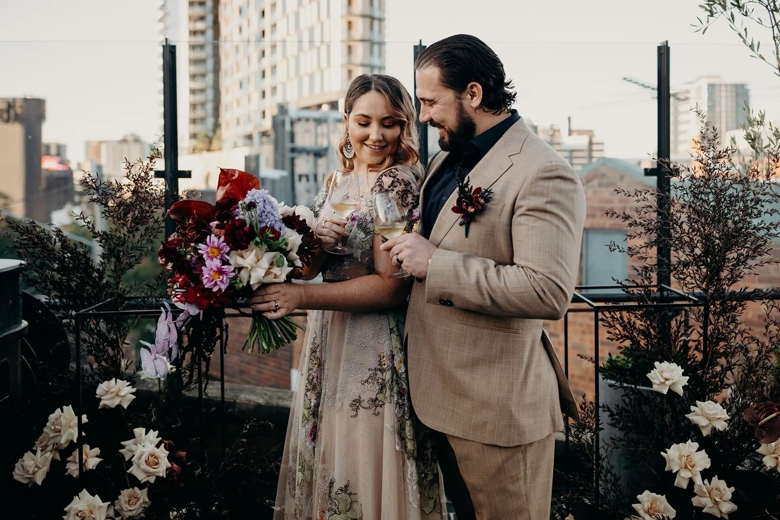 Victoria and Daniel looking down at her colourful bouquet by Mrs Gibbons Flowers on the rooftop terrace, at Agnes Restaurant in Fortitude Valley, Brisbane, for an Elopement Collective elopement. Photo by Tessa Shannon.