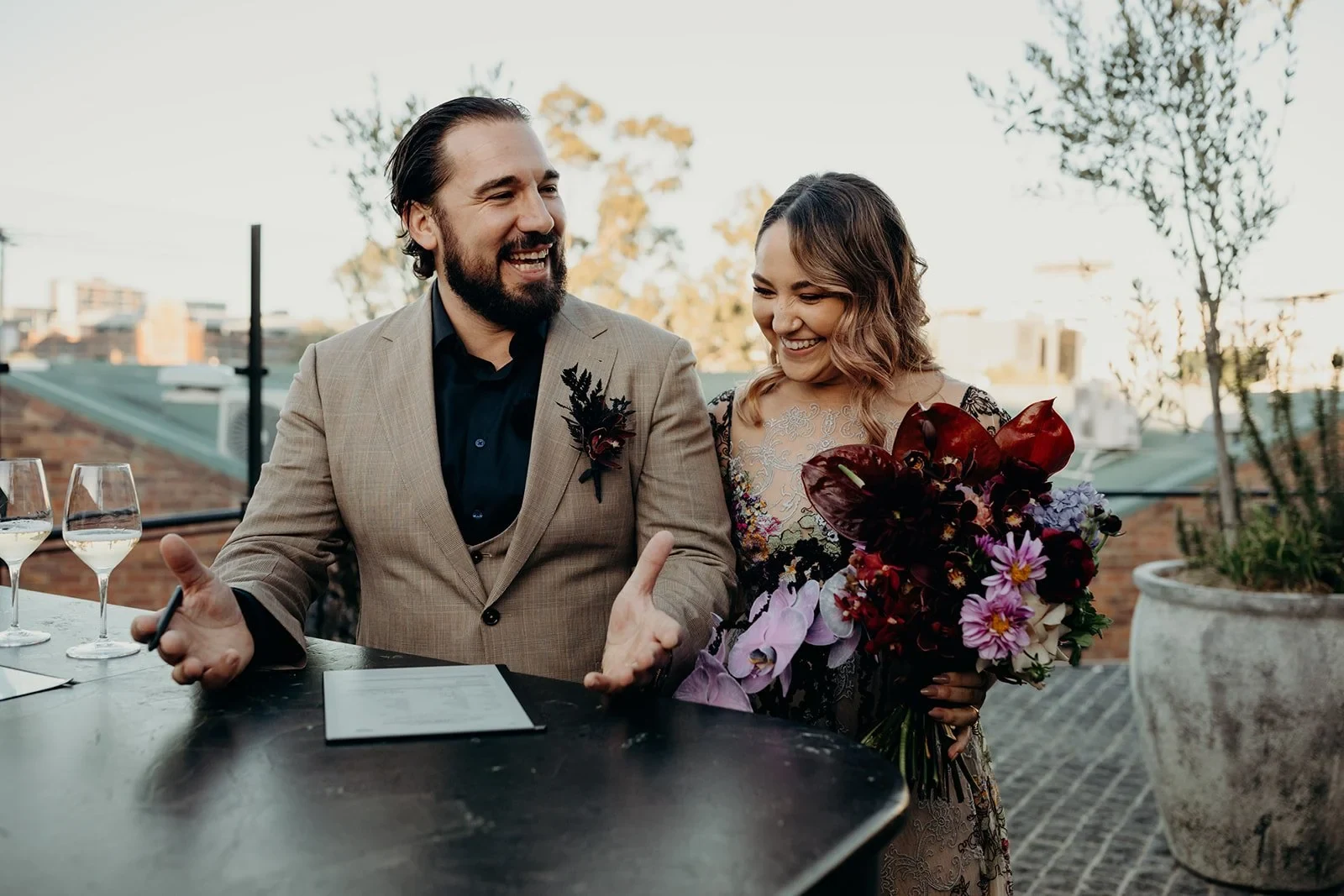 Victoria and Daniel laughing beside their signing paperwork and her bouquet by Mrs Gibbons Flowers, at Agnes Restaurant in Fortitude Valley, Brisbane, for an Elopement Collective elopement. Photo by Tessa Shannon.