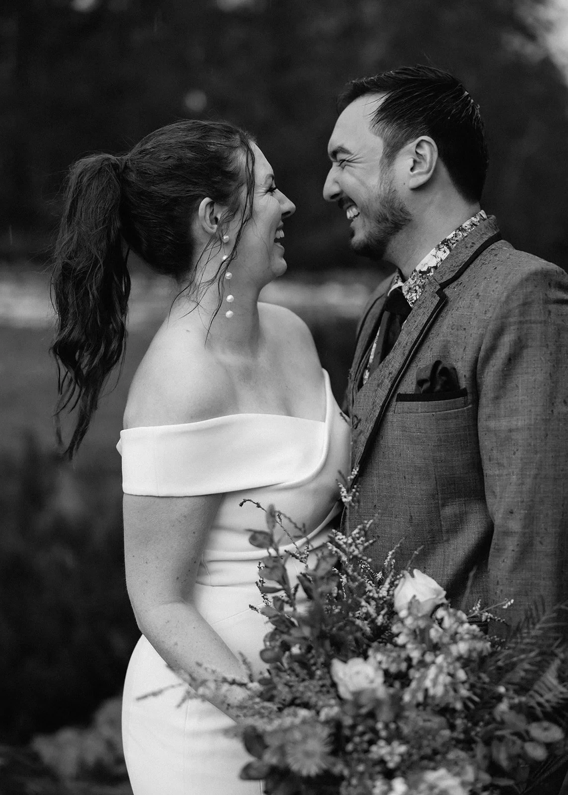 Black-and-white portrait of Sarah and Jono smiling at each other with her bouquet by The Romantics between them, at Pumphouse Point, Lake St Clair, Tasmania, for an Elopement Collective elopement. Photo by Bulb Creative.