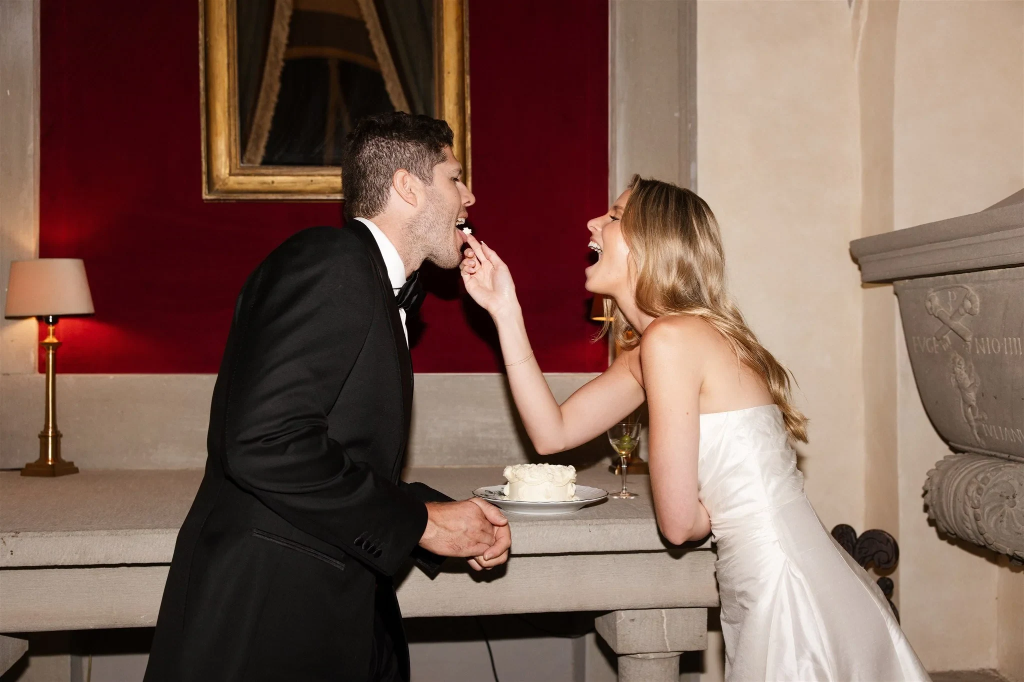 Holly feeding Matt a bite of cake beside a small white cake and a red wall, at Belmond Villa San Michele in Fiesole, overlooking Florence, Tuscany, Italy, for an Elopement Collective elopement. Photo by Khara from House of Lucie.