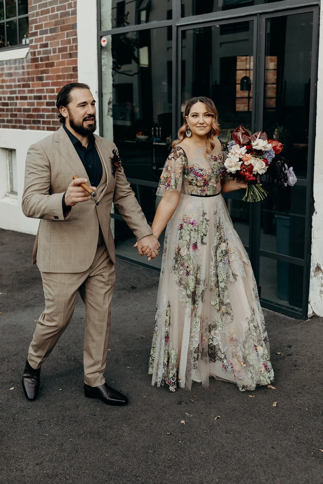 Full-length portrait of Victoria and Daniel holding hands outside a dark doorway, with her bouquet by Mrs Gibbons Flowers, in Fortitude Valley, Brisbane, for an Elopement Collective elopement. Photo by Tessa Shannon.