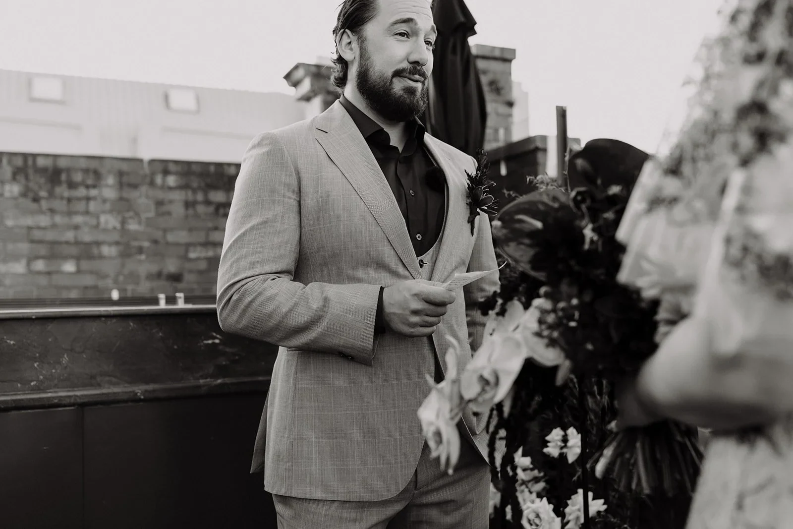 Black-and-white photograph of Daniel reading his vows to Victoria on the rooftop terrace, at Agnes Restaurant in Fortitude Valley, Brisbane, for an Elopement Collective elopement. Photo by Tessa Shannon.