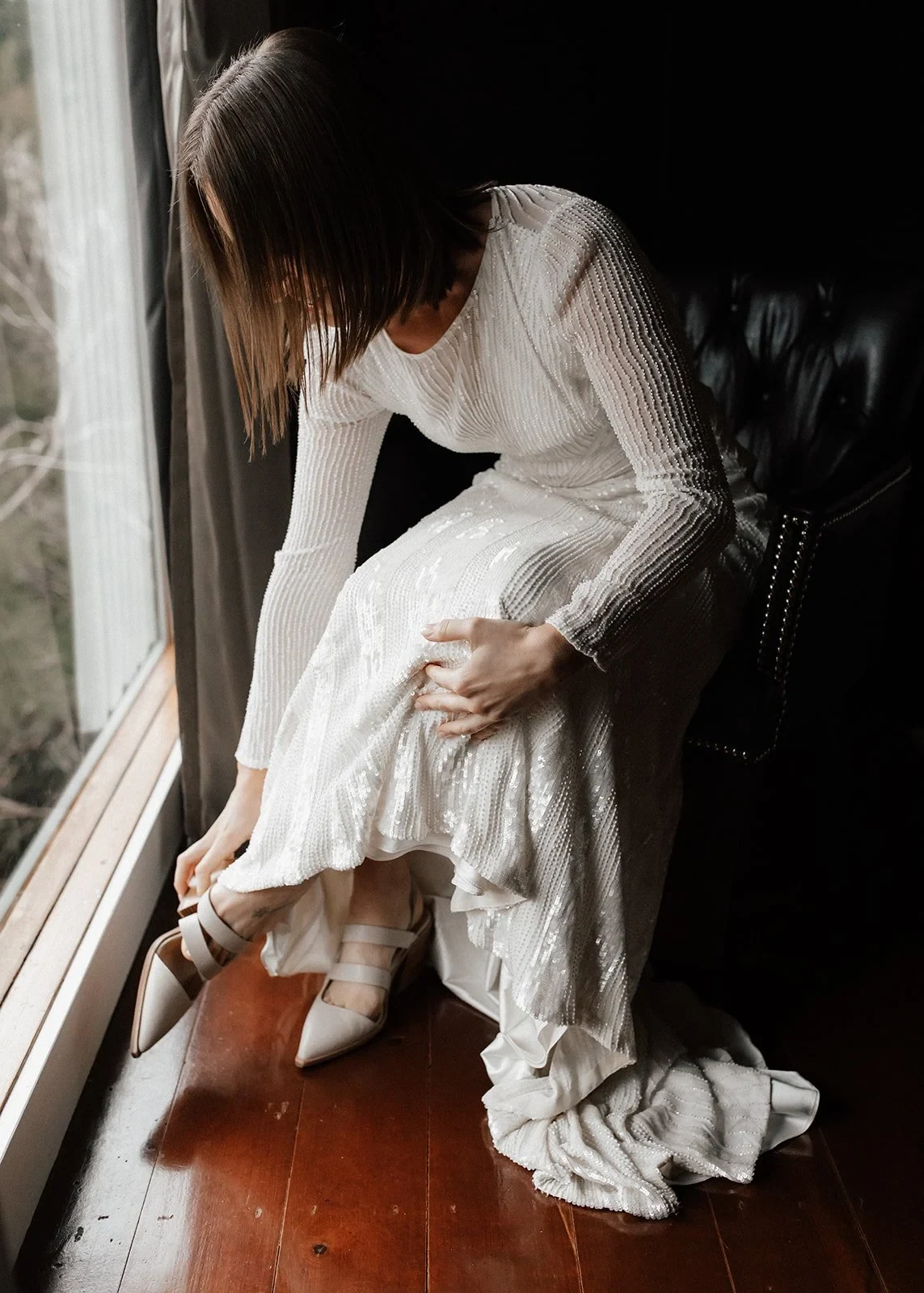 April lifting the skirt of her Karen Willis Holmes dress to fasten a white heel beside the window, at Cradle Mountain Lodge in Cradle Mountain, Tasmania, for an Elopement Collective elopement. Photo by Bulb Creative.