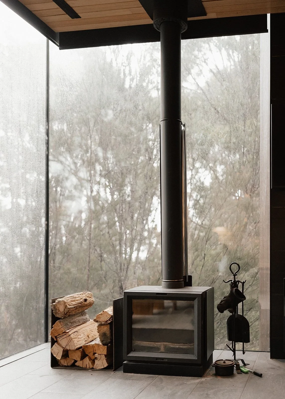 A wood-burning stove and stacked logs beside floor-to-ceiling windows looking into the trees, at Pumphouse Point, Lake St Clair, Tasmania, for an Elopement Collective elopement. Photo by Bulb Creative.