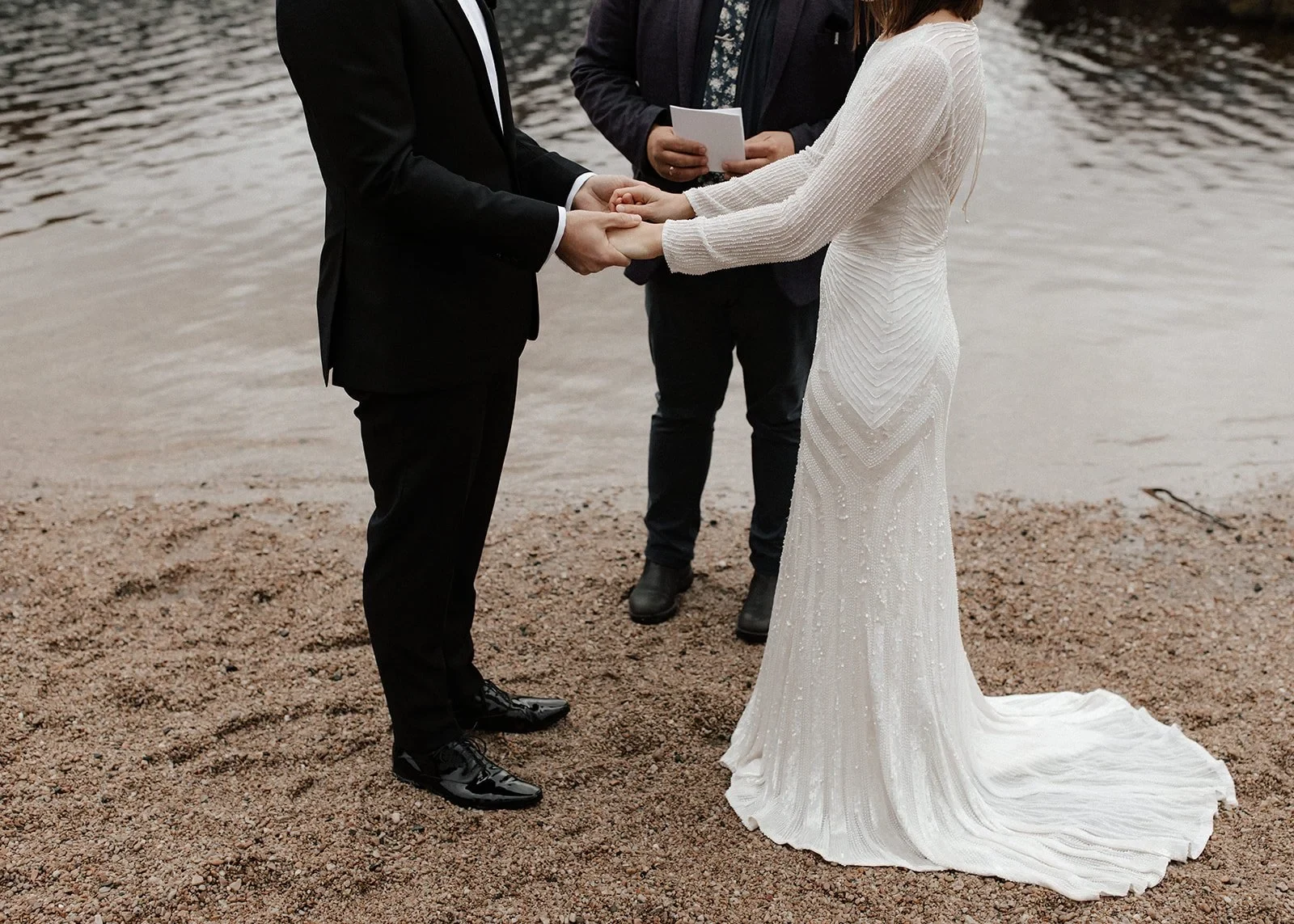 Close view of April and Josh holding hands, with her white dress trailing over the sand and Josh Withers behind them, at Dove Lake, Cradle Mountain, Tasmania, for an Elopement Collective elopement. Photo by Bulb Creative.