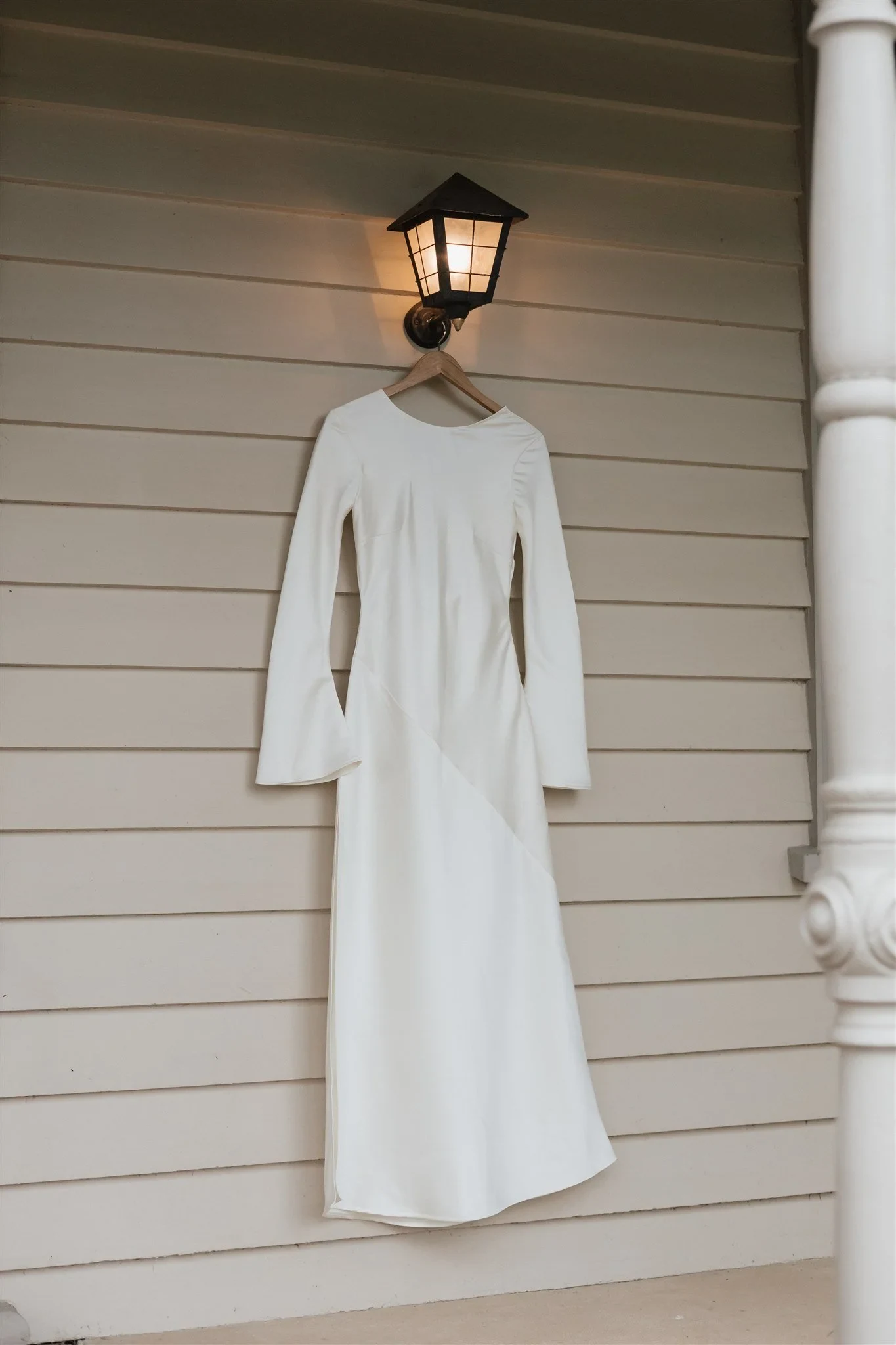 Maddy’s white Olivia Rose the Label dress hanging on a weatherboard veranda beneath a lantern, at The Mayor’s Residence in Daylesford, Victoria, for an Elopement Collective elopement. Photo by Bulb Creative.