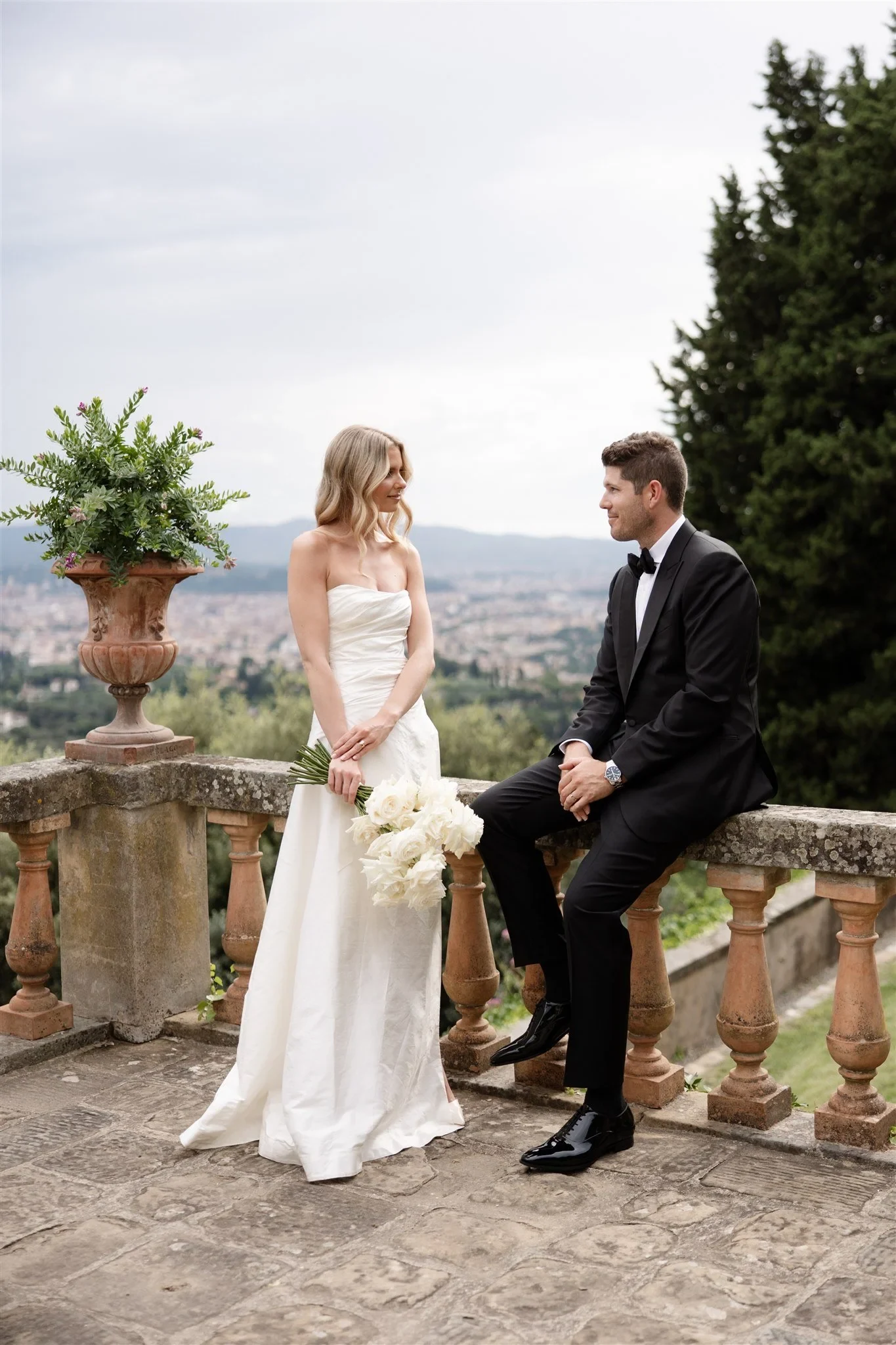 Holly holding her white bouquet by Le Francesche beside Matt as he sits on the stone balustrade, at Belmond Villa San Michele in Fiesole, overlooking Florence, Tuscany, Italy, for an Elopement Collective elopement. Photo by Khara from House of Lucie.