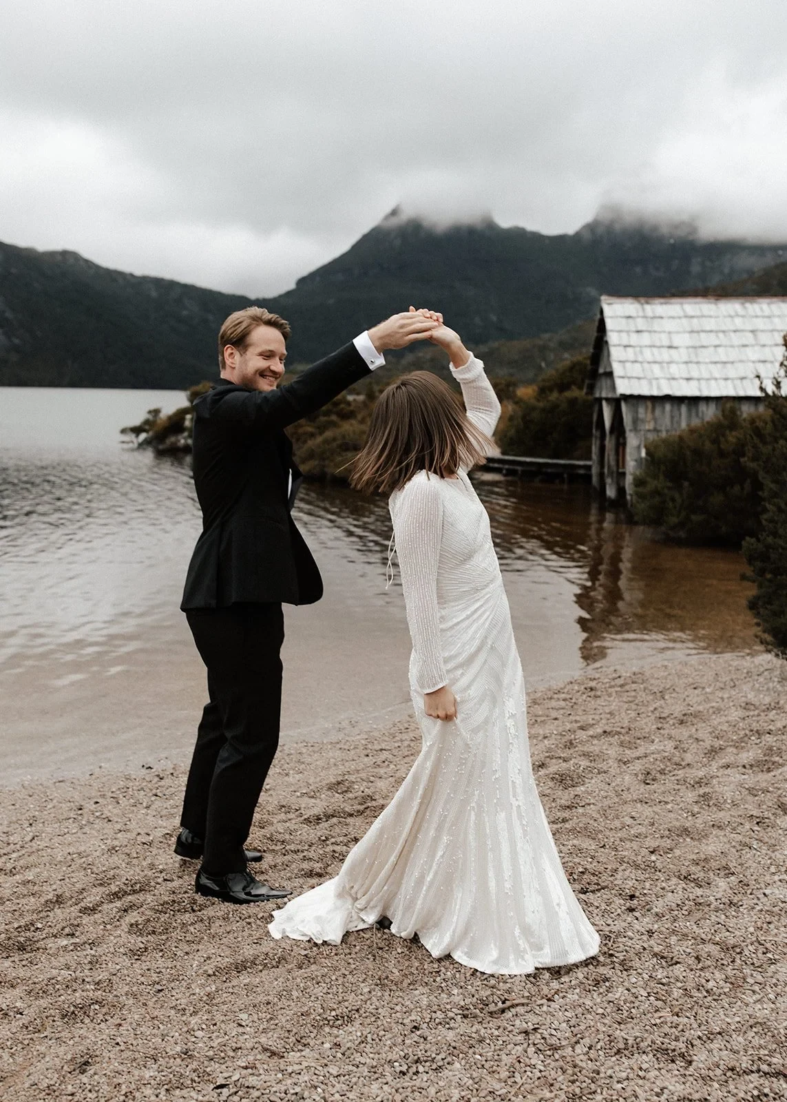 Josh twirling April on the sand beside the stone boatshed, at Dove Lake, Cradle Mountain, Tasmania, for an Elopement Collective elopement. Photo by Bulb Creative.