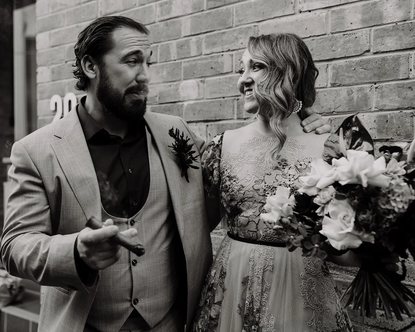 Black-and-white portrait of Daniel with a cigar beside Victoria and her bouquet by Mrs Gibbons Flowers, in Fortitude Valley, Brisbane, for an Elopement Collective elopement. Photo by Tessa Shannon.