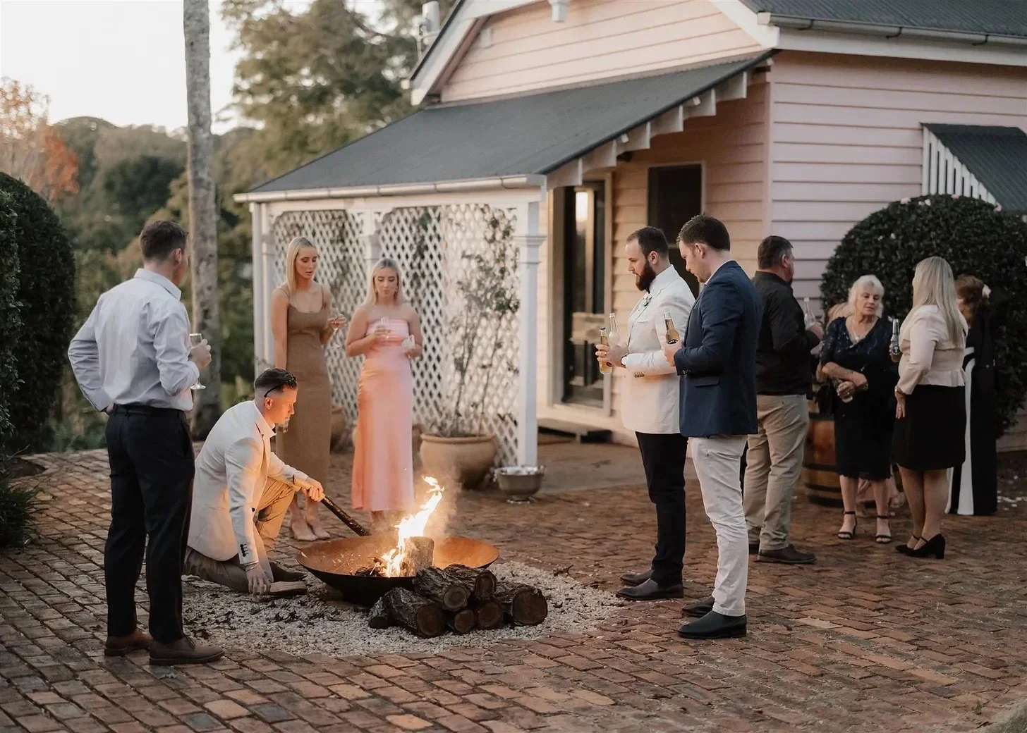 Guests gathering beside a courtyard fire outside a pale weatherboard house, in Montville, Sunshine Coast Hinterland, Queensland, for an Elopement Collective elopement. Photo by Bulb Creative.