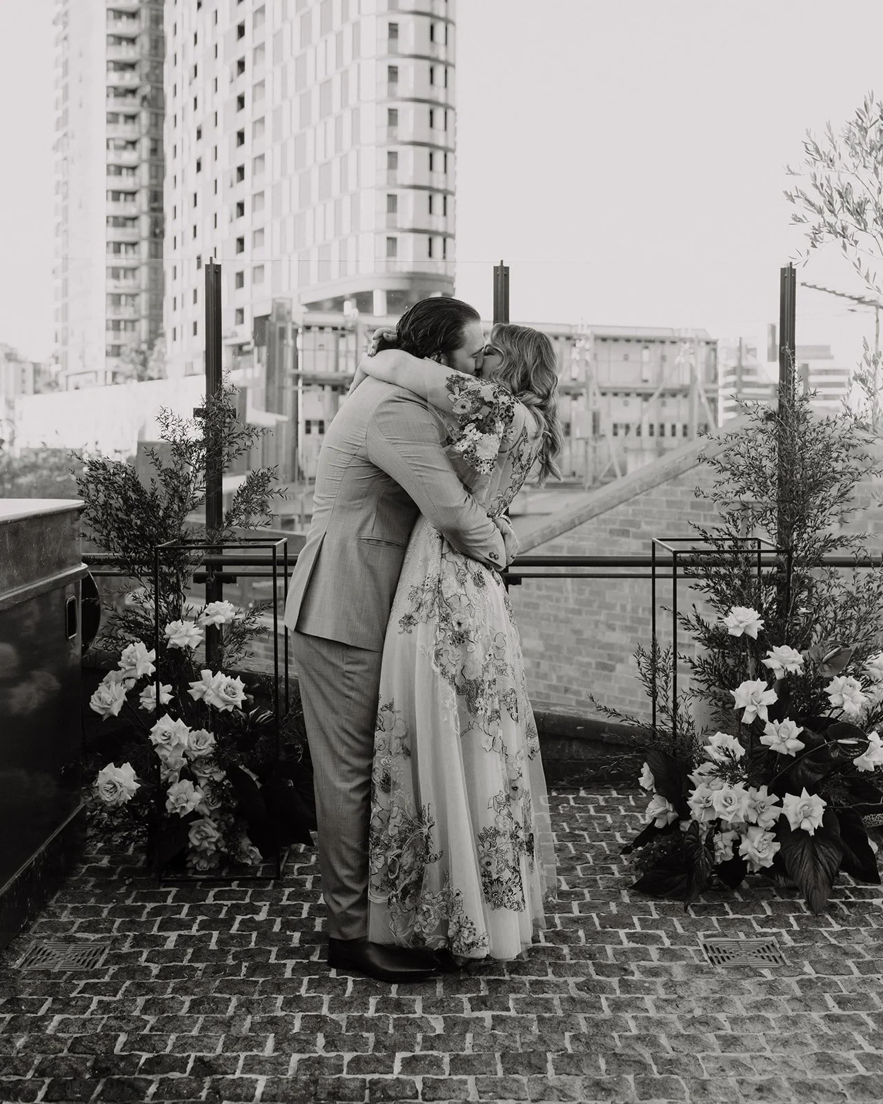 Black-and-white photograph of Victoria and Daniel kissing between ceremony flowers by Mrs Gibbons Flowers, at Agnes Restaurant in Fortitude Valley, Brisbane, for an Elopement Collective elopement. Photo by Tessa Shannon.