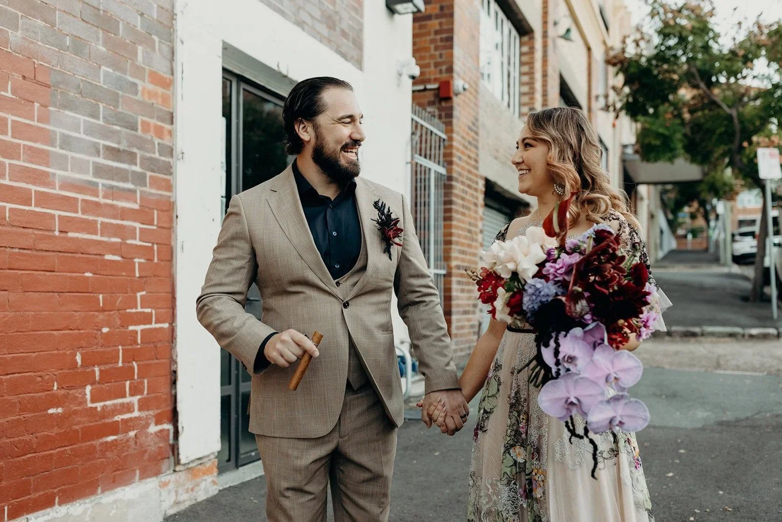 Victoria and Daniel laughing and holding hands beside a red brick building, with her bouquet by Mrs Gibbons Flowers, in Fortitude Valley, Brisbane, for an Elopement Collective elopement. Photo by Tessa Shannon.