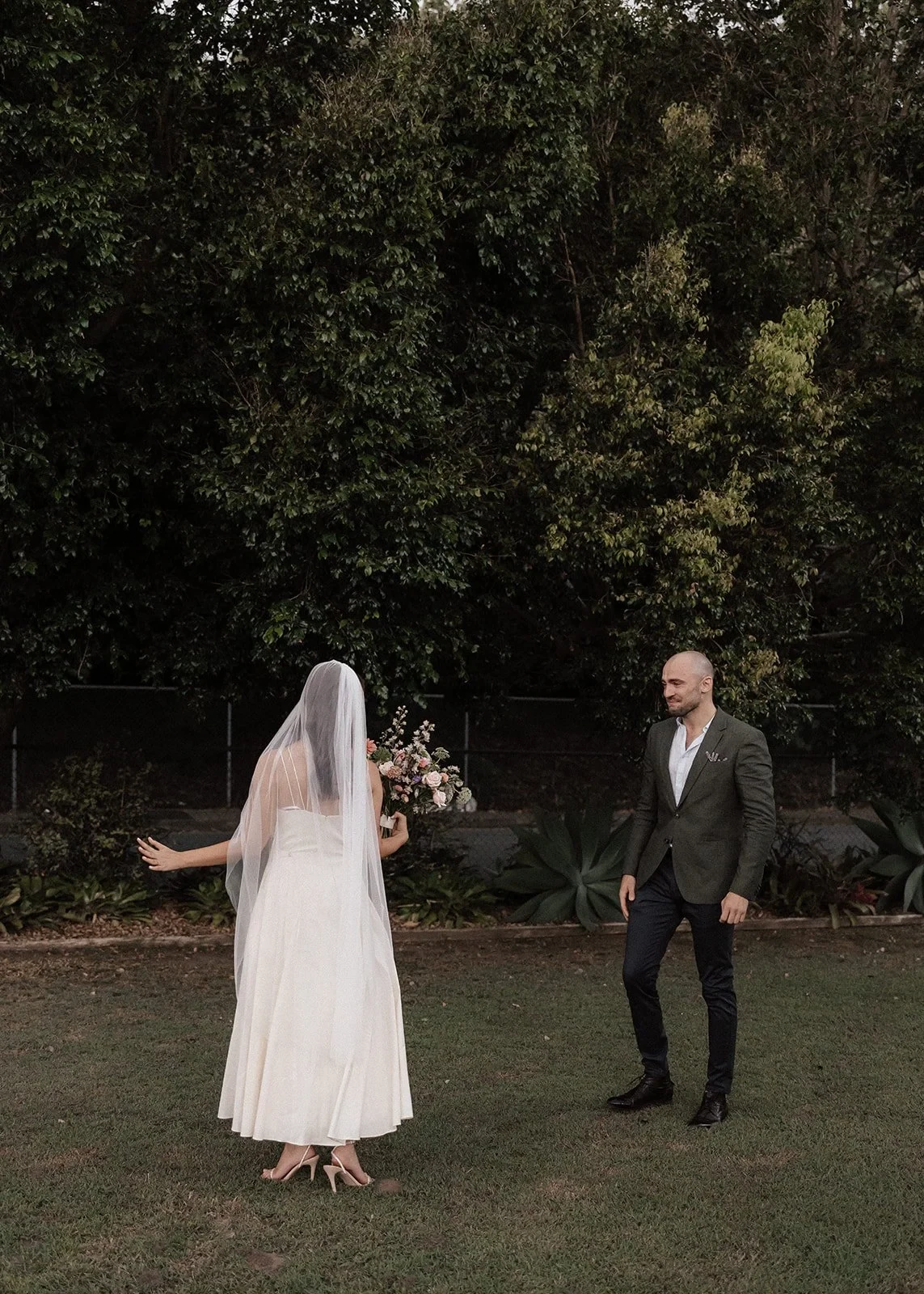 Veiled bride holding a bouquet faces her partner on a garden lawn