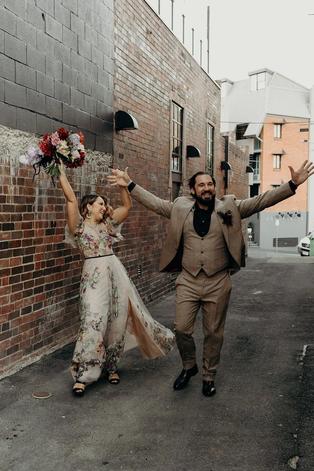 Victoria and Daniel running through a laneway with their arms raised and her bouquet by Mrs Gibbons Flowers held aloft, in Fortitude Valley, Brisbane, for an Elopement Collective elopement. Photo by Tessa Shannon.