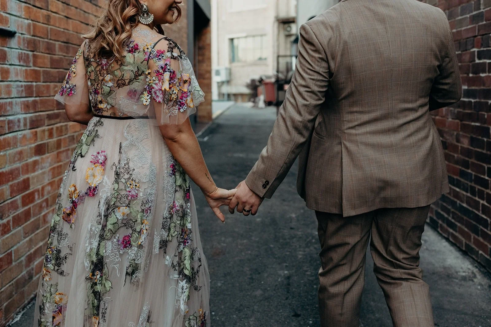 View from behind of Victoria and Daniel holding hands in a narrow brick-lined laneway, in Fortitude Valley, Brisbane, for an Elopement Collective elopement. Photo by Tessa Shannon.