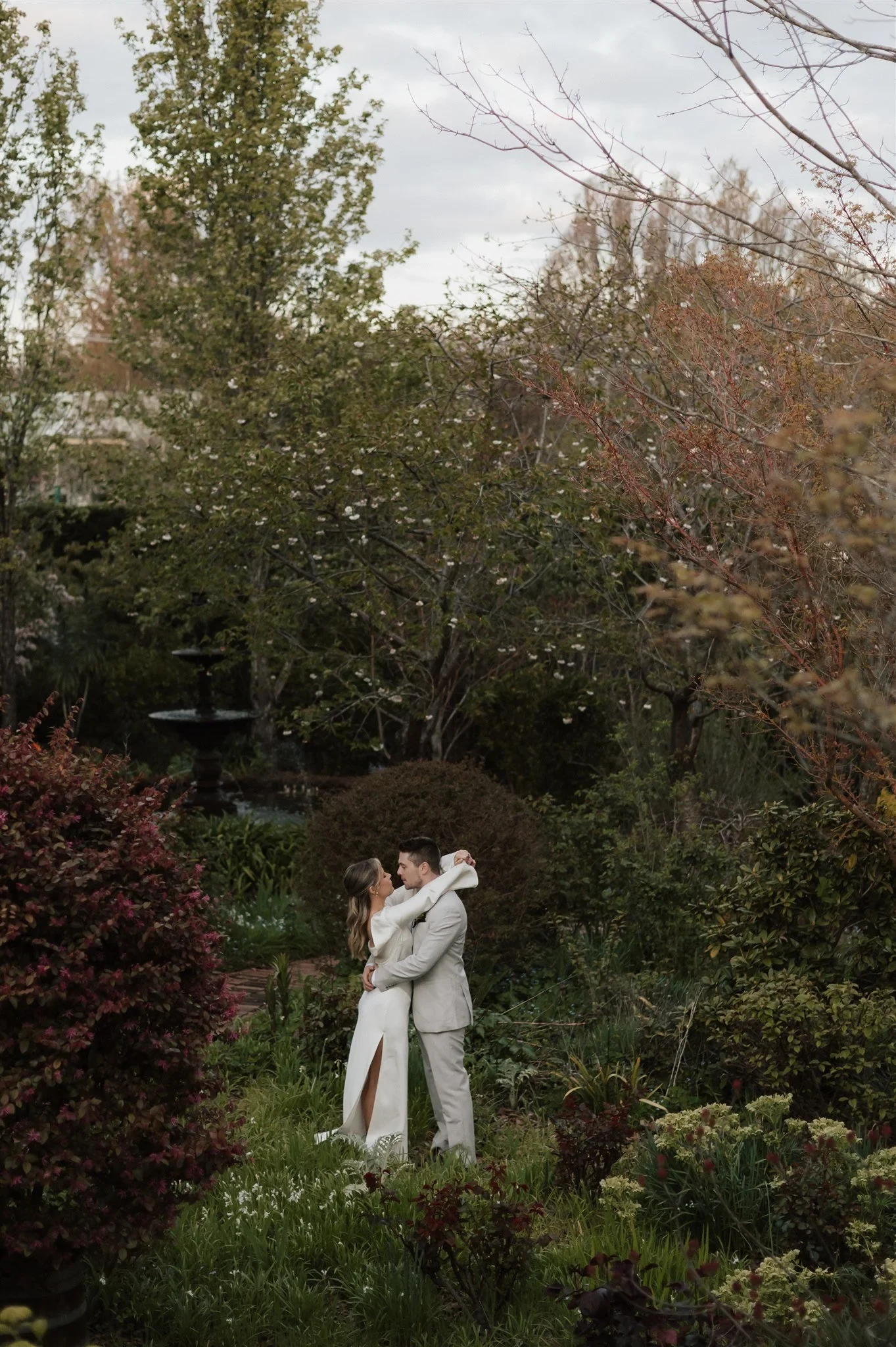 Maddy and Cameron embracing among flowering garden beds and autumn trees, at The Mayor’s Residence in Daylesford, Victoria, for an Elopement Collective elopement. Photo by Bulb Creative.