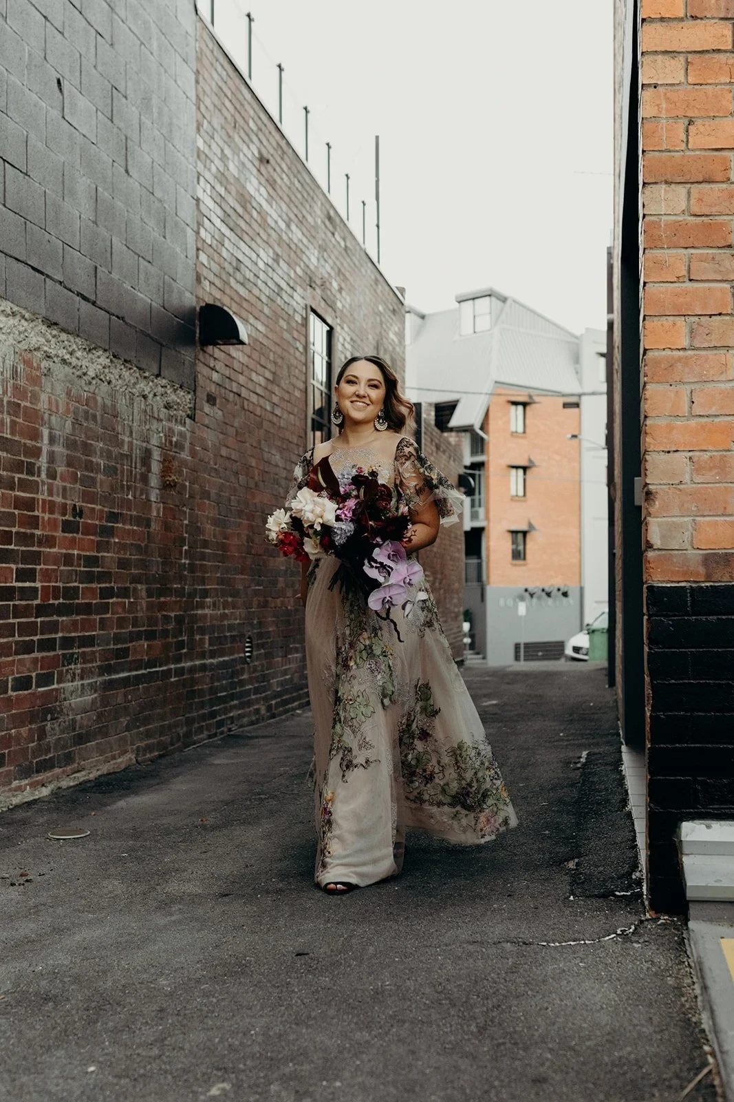 Full-length portrait of Victoria with her bouquet by Mrs Gibbons Flowers in a narrow brick-lined laneway, in Fortitude Valley, Brisbane, for an Elopement Collective elopement. Photo by Tessa Shannon.