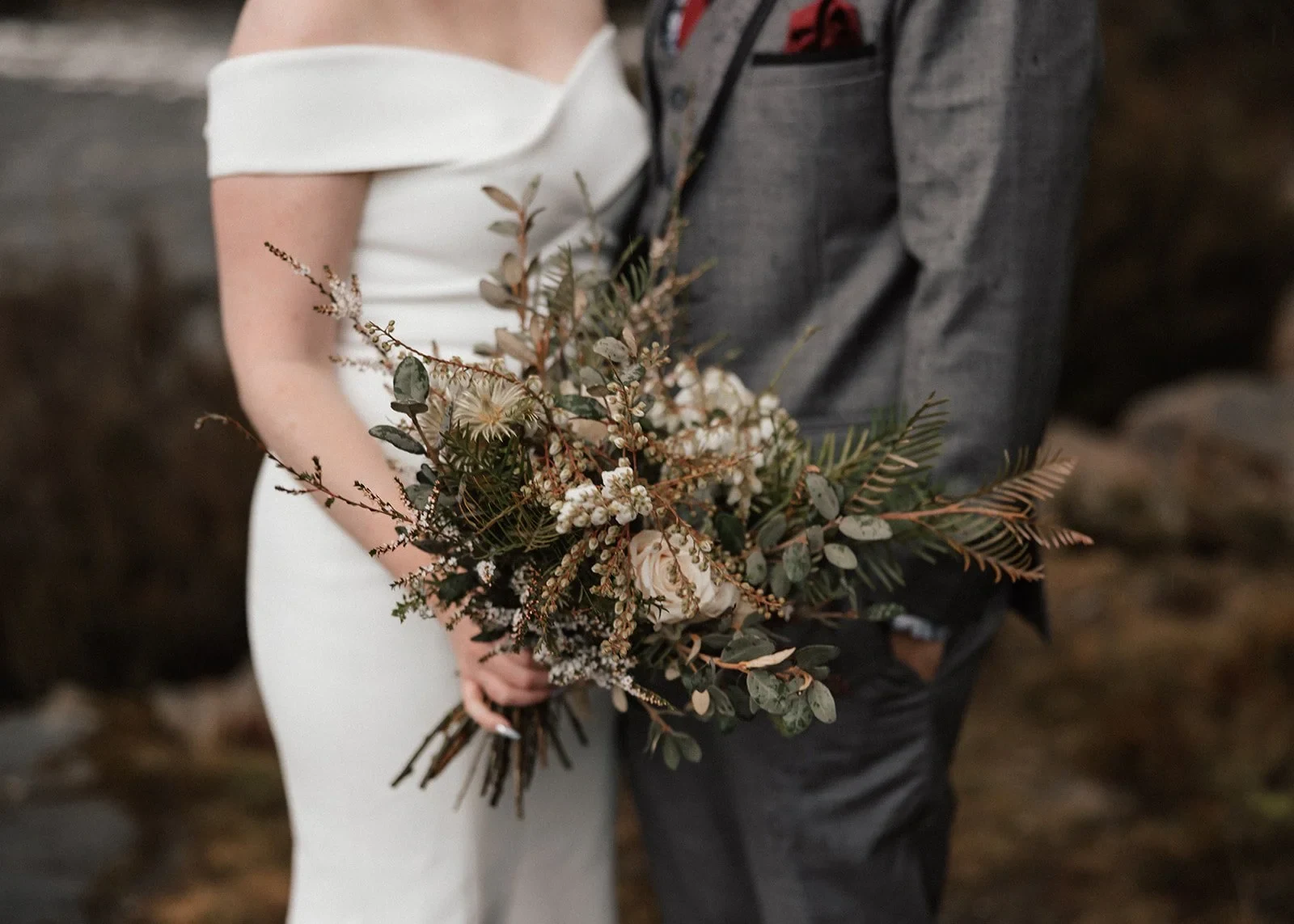 Sarah holding her bouquet of muted pink and cream flowers and foliage by The Romantics beside Jono’s grey suit, at Pumphouse Point, Lake St Clair, Tasmania, for an Elopement Collective elopement. Photo by Bulb Creative.