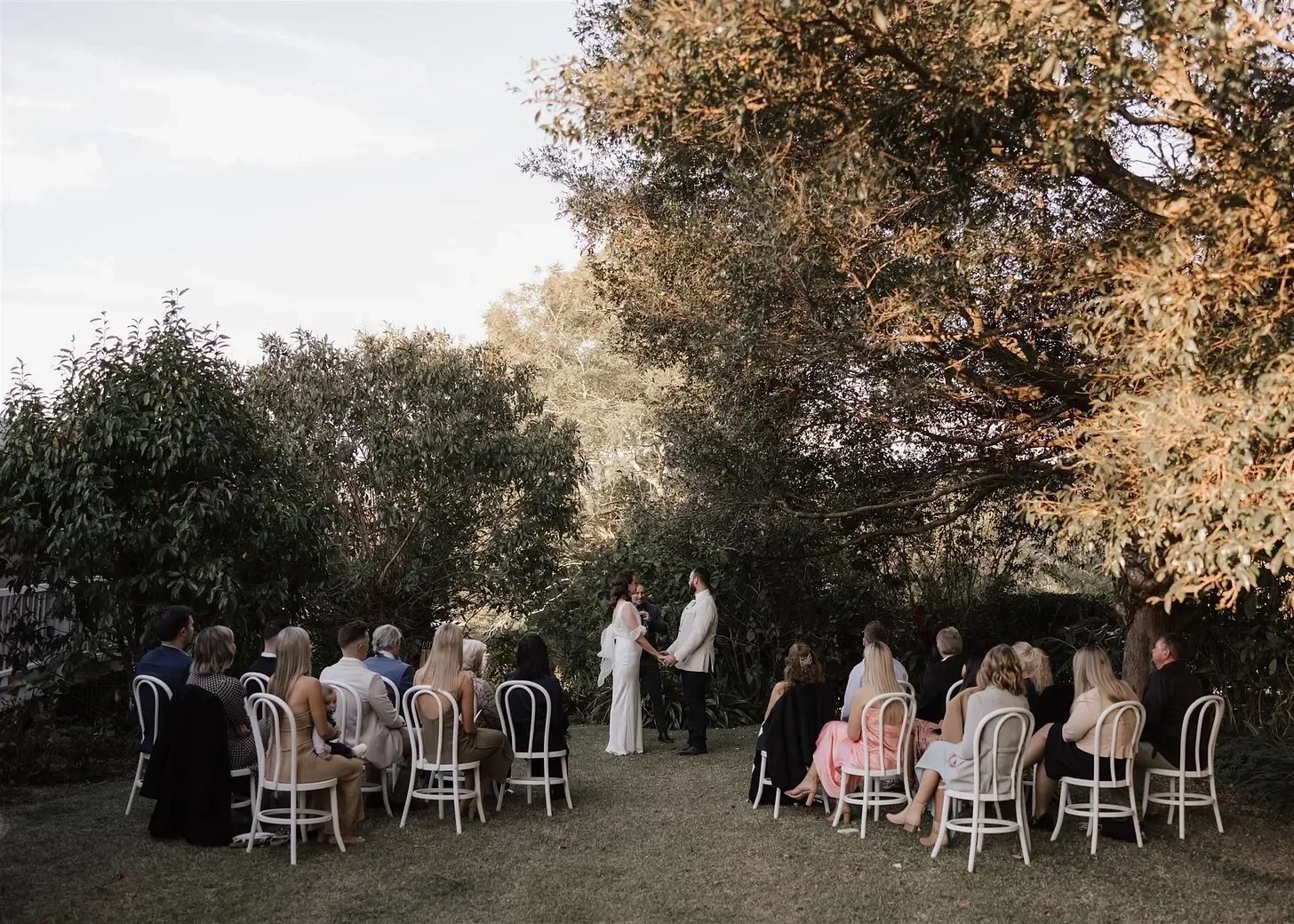 Tineeka and Chris standing with celebrant Jake Smith beneath garden trees, with guests seated nearby, in Montville, Sunshine Coast Hinterland, Queensland, for an Elopement Collective elopement. Photo by Bulb Creative.