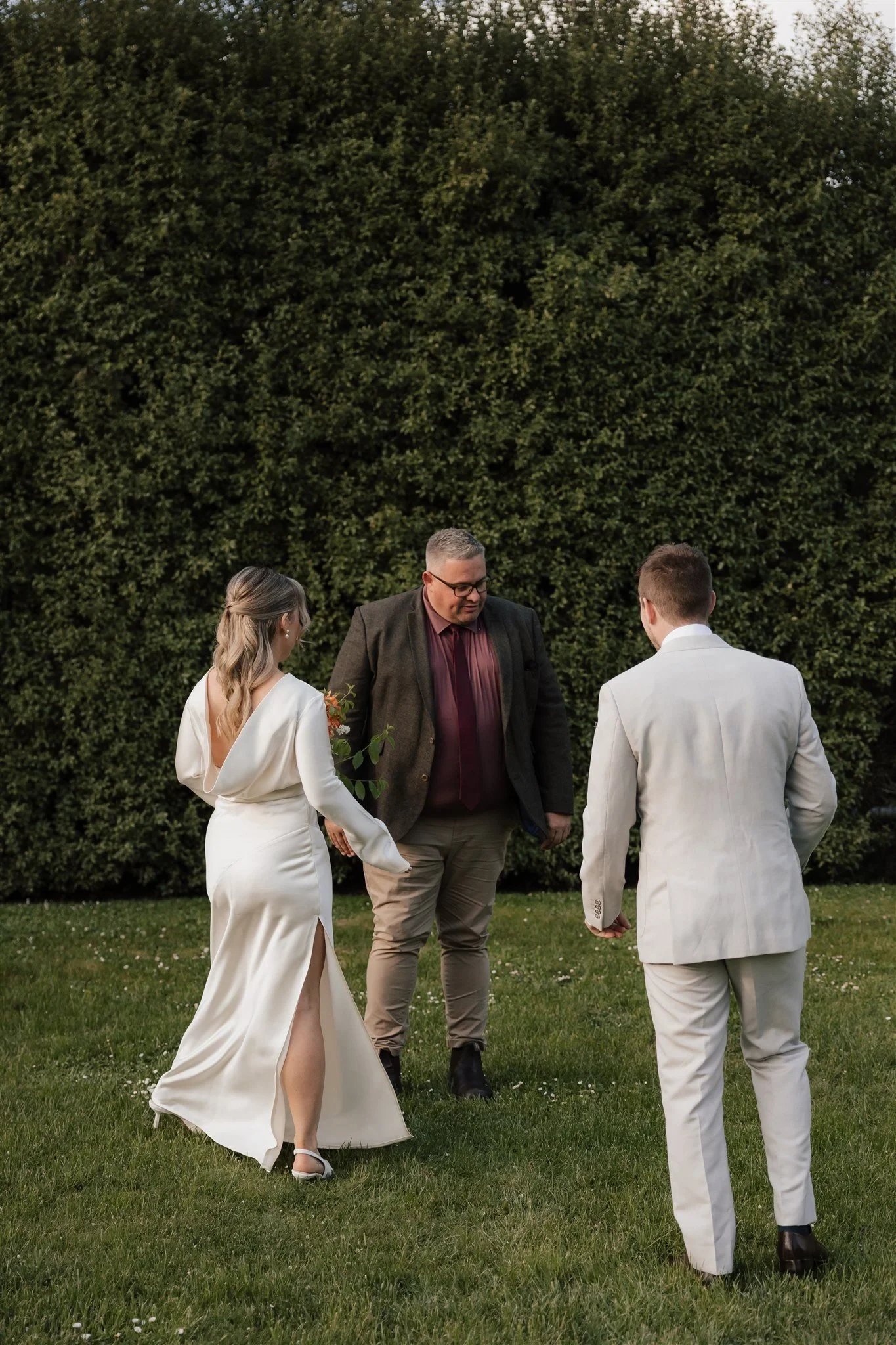Maddy raising her hand to her face as she and Cameron stand with celebrant Josh Withers on the garden lawn, at The Mayor’s Residence in Daylesford, Victoria, for an Elopement Collective elopement. Photo by Bulb Creative.