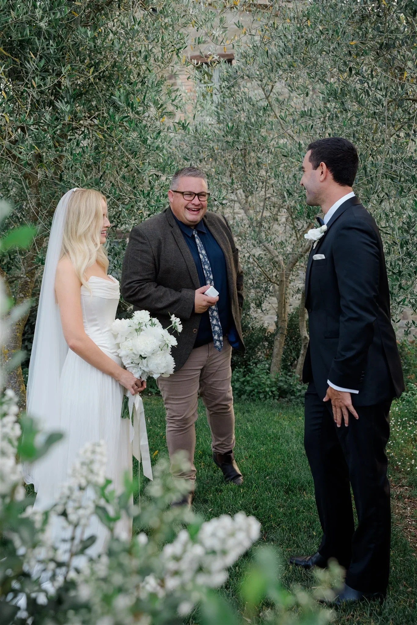 Shelby and Jack listening to celebrant Josh Withers beneath leafy garden trees, in Umbria, Italy, for an Elopement Collective elopement. Photo by James from House of Love.