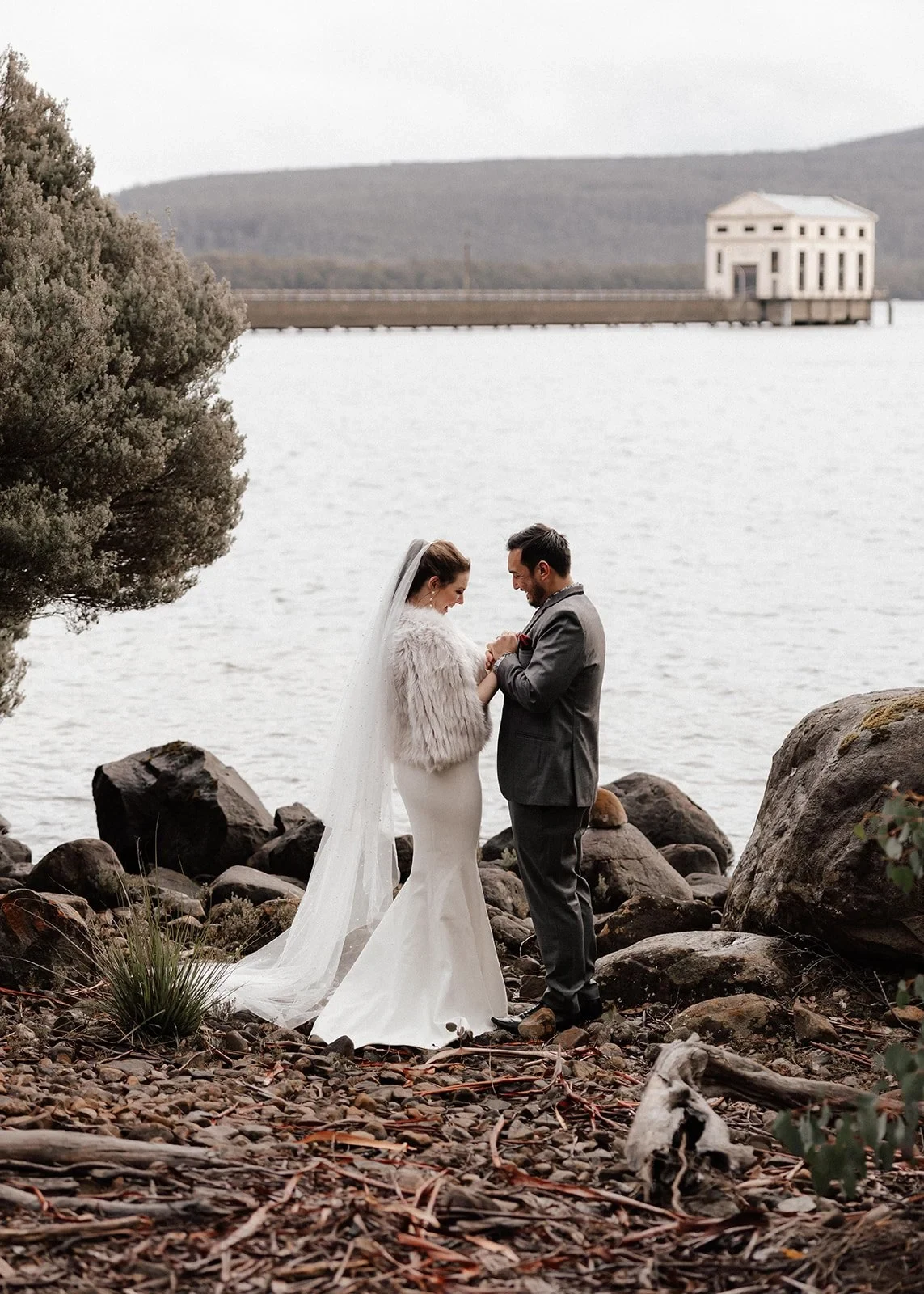 Sarah and Jono standing close together among lakeside rocks, with the Pumphouse building in the distance, at Pumphouse Point, Lake St Clair, Tasmania, for an Elopement Collective elopement. Photo by Bulb Creative.