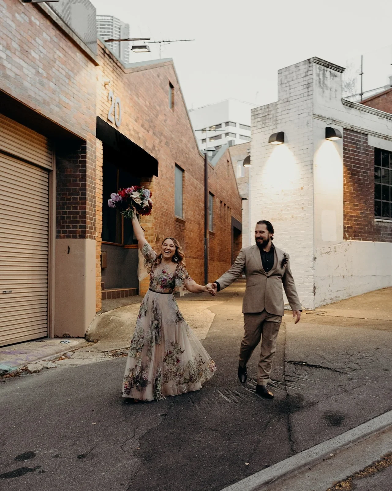Wide view of Victoria and Daniel celebrating while walking hand in hand past a brick building, her bouquet by Mrs Gibbons Flowers raised, in Fortitude Valley, Brisbane, for an Elopement Collective elopement. Photo by Tessa Shannon.